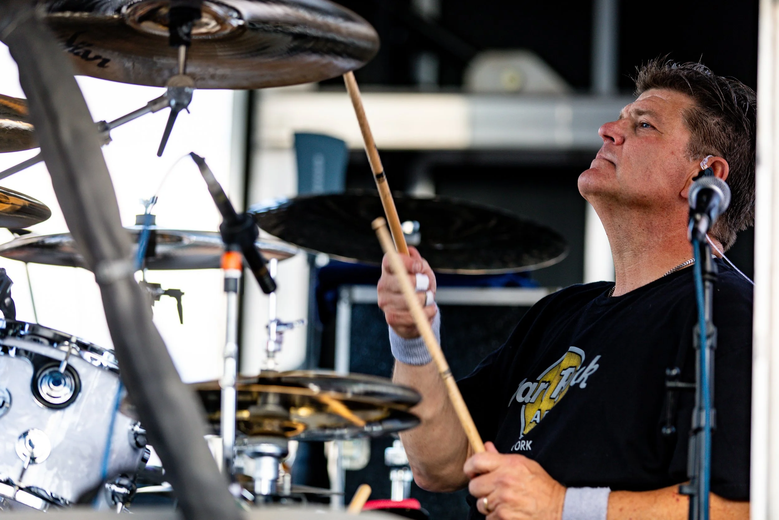 Male drummer playing drums with drumsticks, wearing a black T-shirt, in a performance setting.