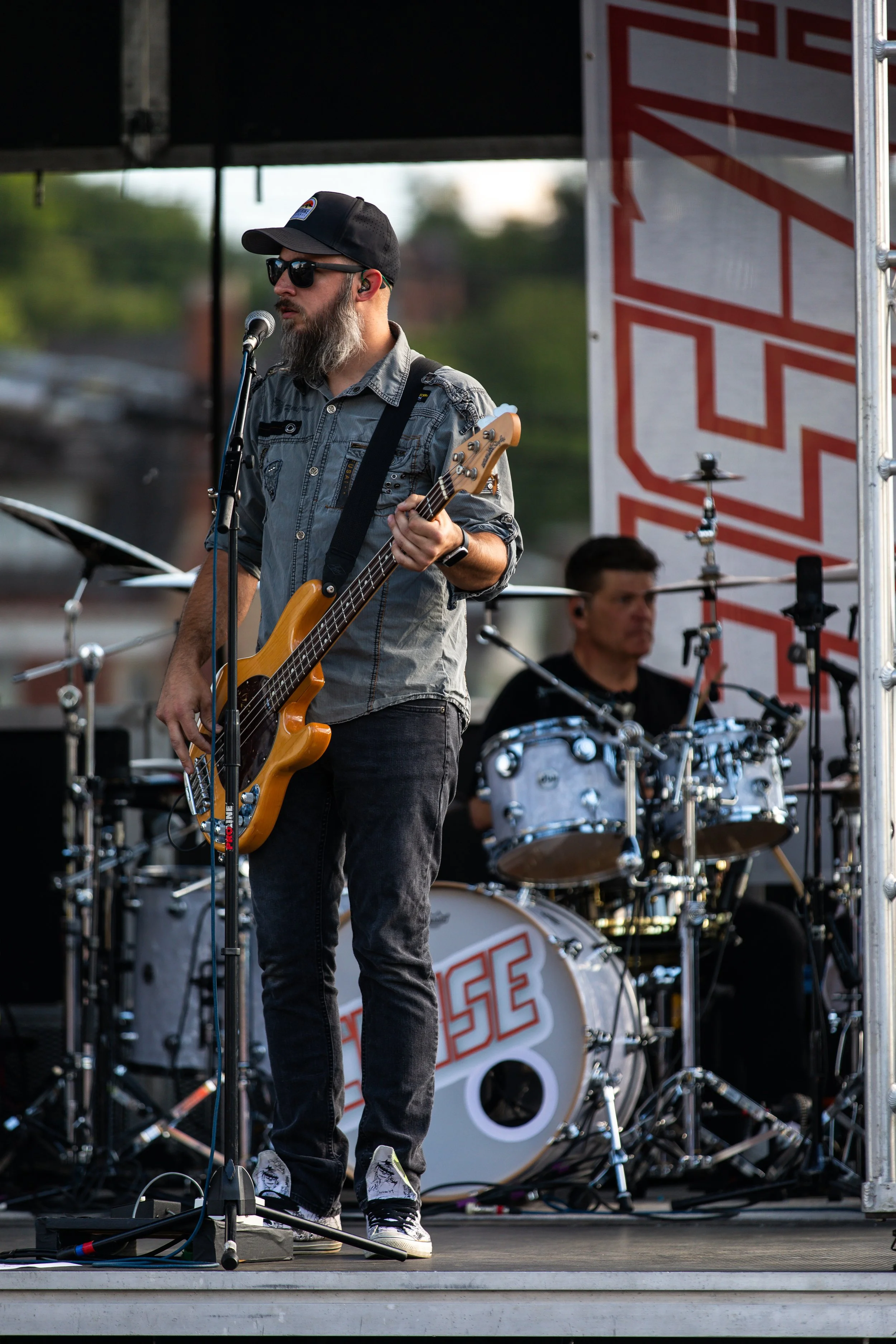 A musician with a beard, sunglasses, and a baseball cap playing an electric bass on stage; a drummer behind him is playing a drum set with the band name 'The Fuse' on the bass drum.