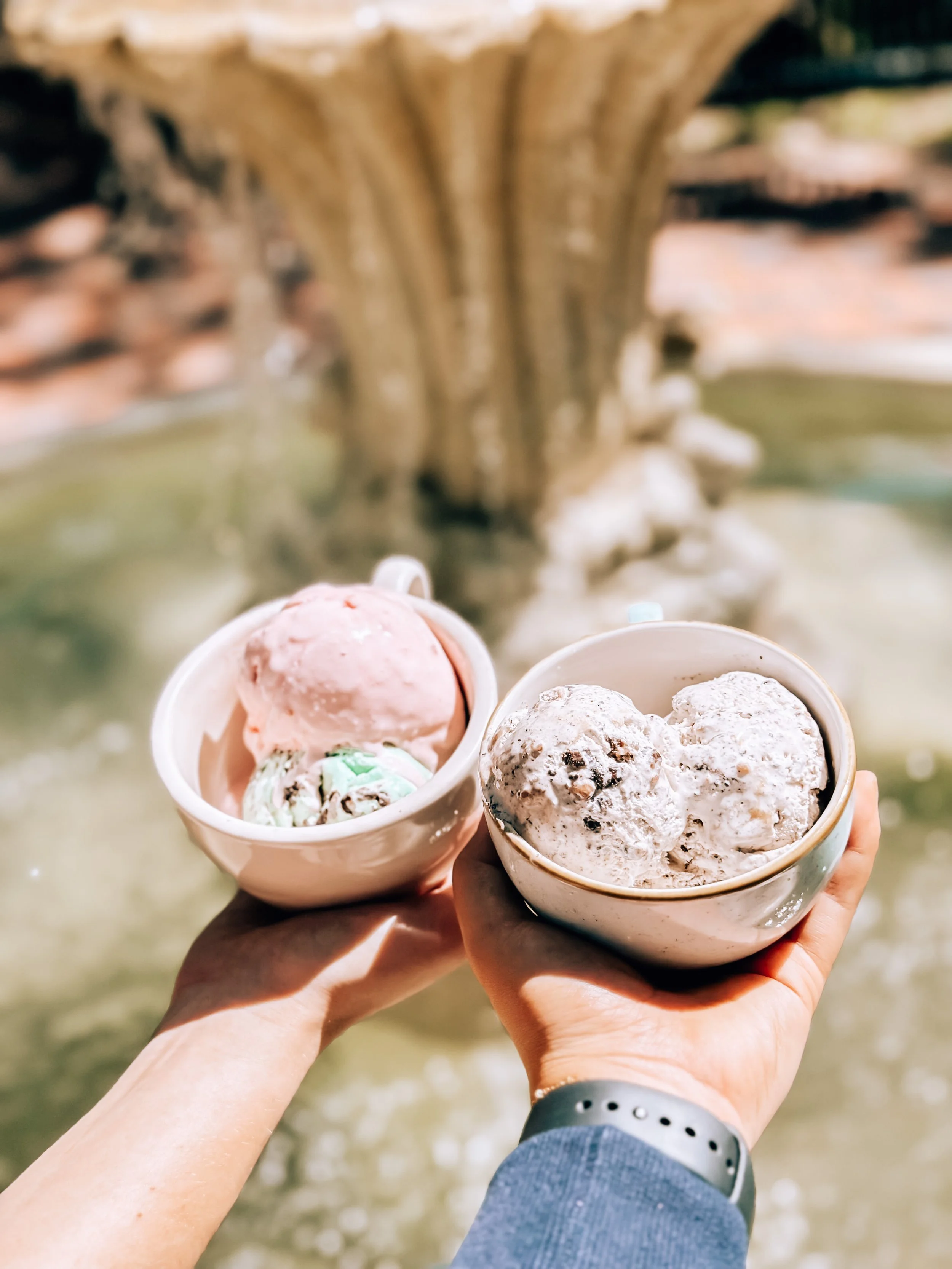 Two hands are side by side each holding a coffee cup with two scoops of ice cream, overlooking the fountain in the Haile Plantation Village Center