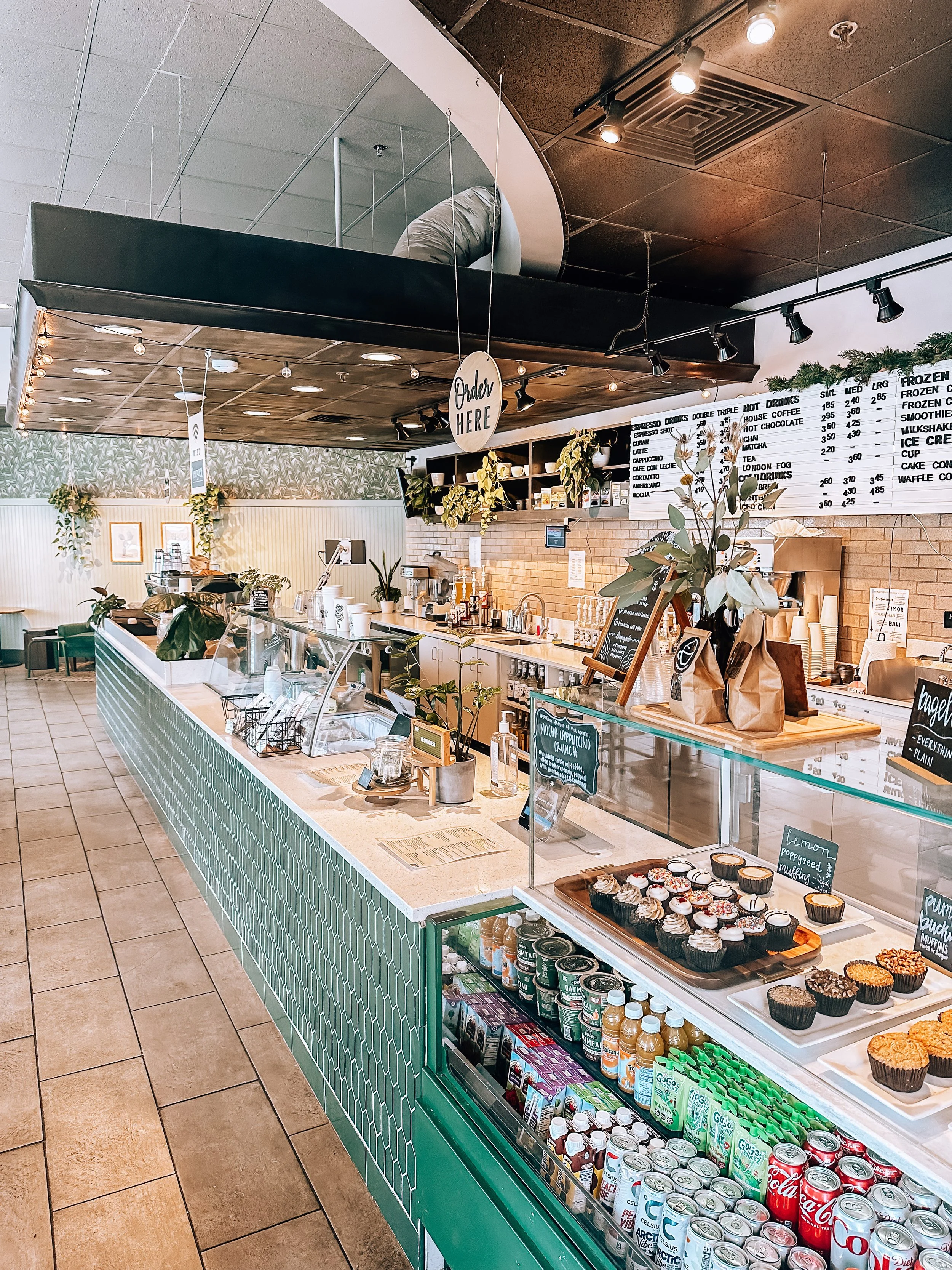 A coffee counter with a display case of coffee bags, assorted cupcakes and muffins, decorated with plants and a large menu board at the back