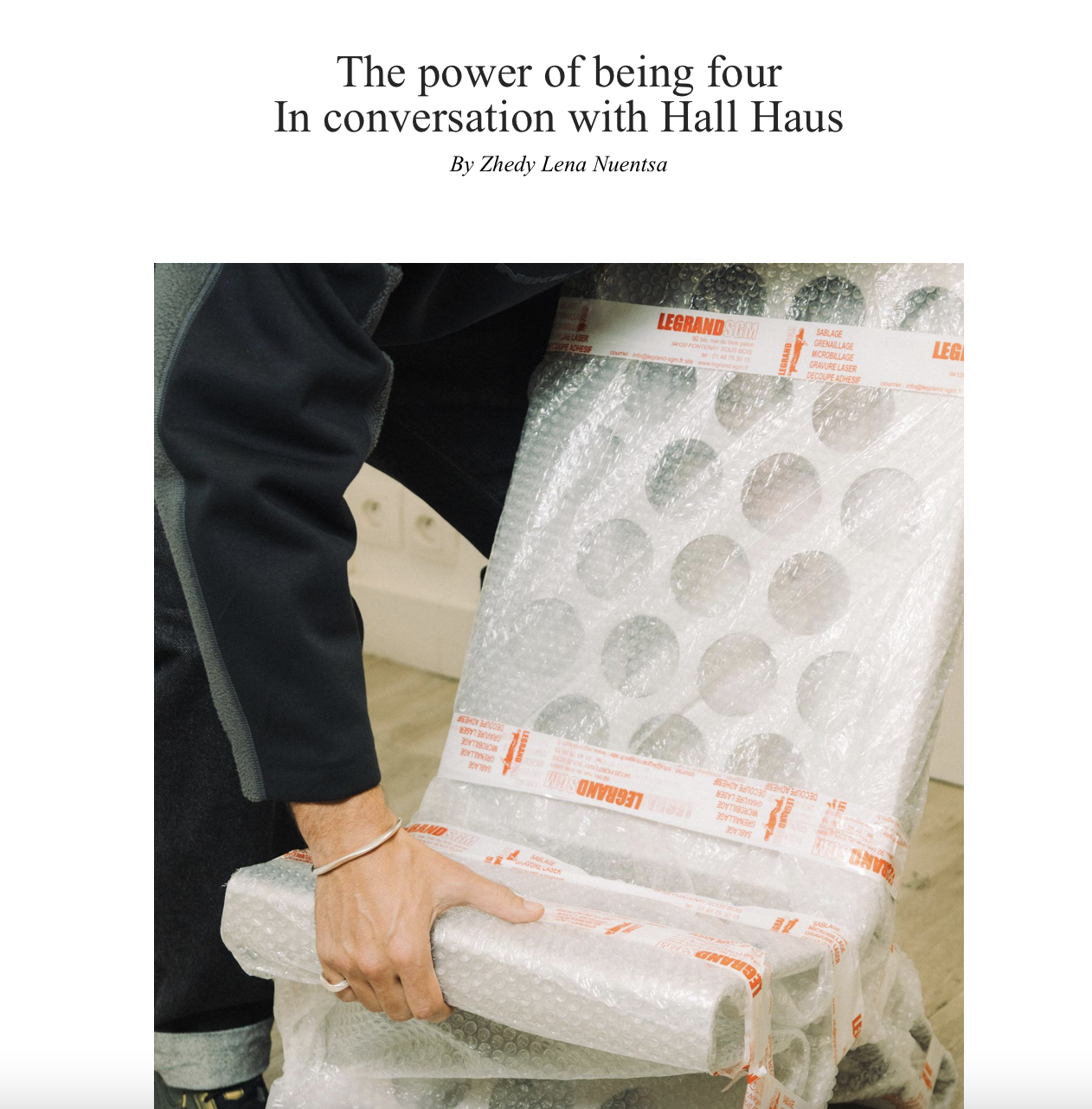 Une personne portant un vêtement noir, tenant un matelas ou un objet emballé dans du plastique à bulles, avec des bandes en plastique imprimées en orange