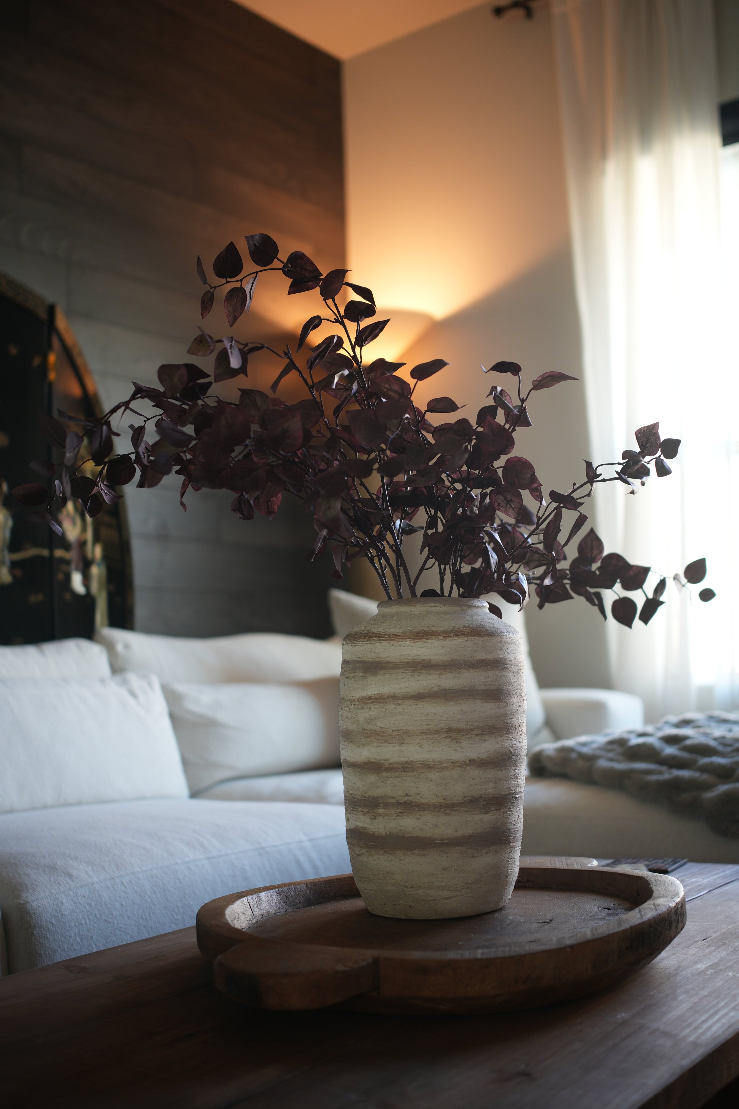 A beige ceramic vase with dark red leaves on a wooden tray in a cozy living room with a white sofa and natural light from a window.