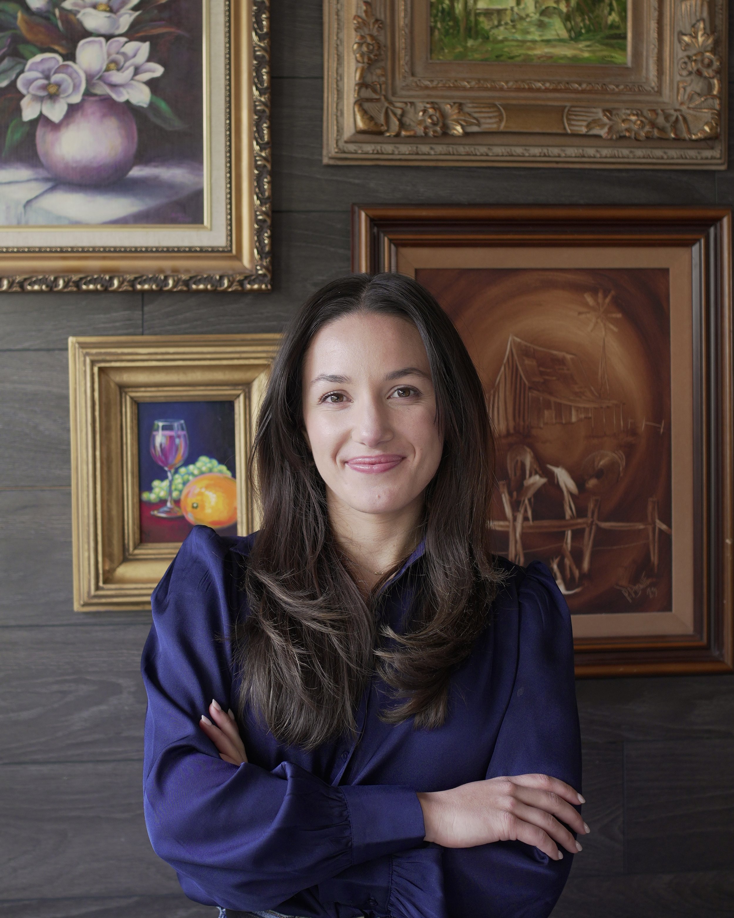 A woman with long dark hair wearing a navy blue blouse standing in front of a wall decorated with several framed paintings.