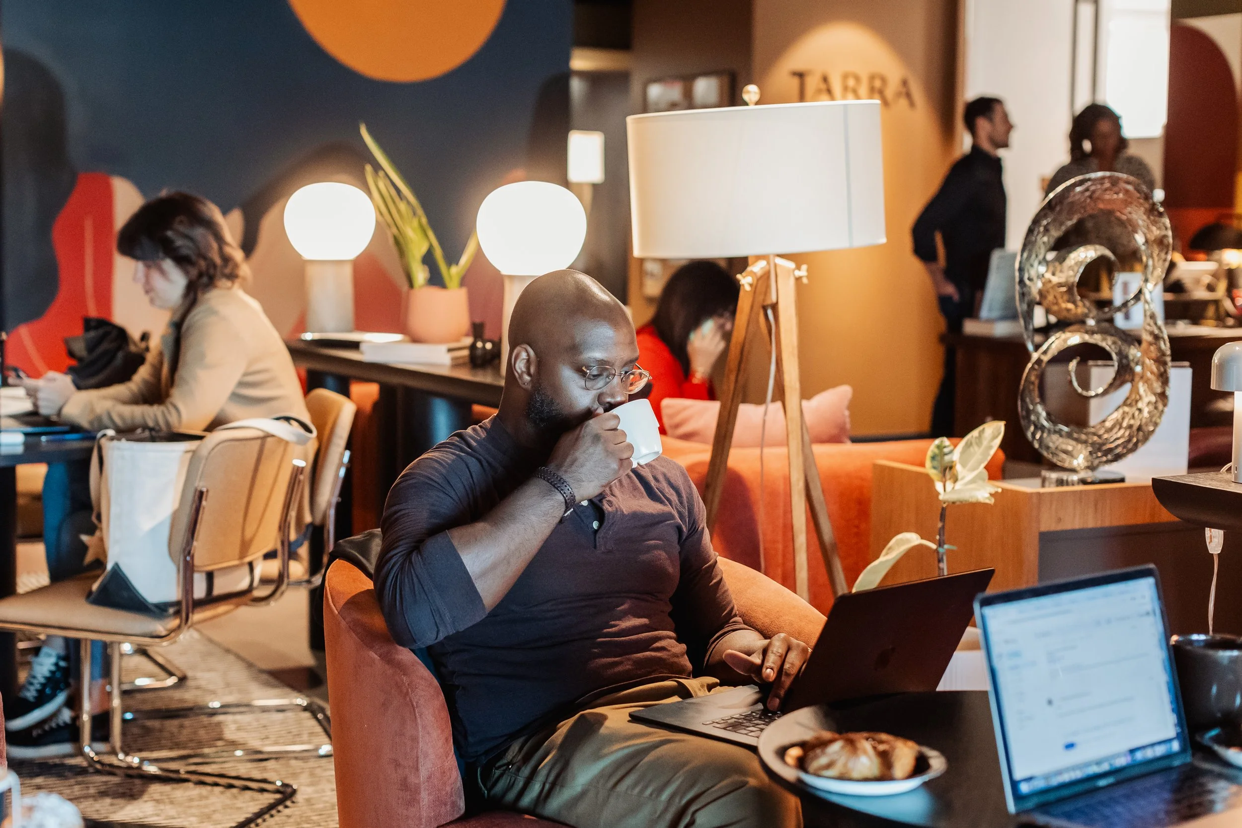 A man sitting in a cozy cafe, wearing glasses, drinking from a white mug while working on a laptop, with a plate of croissants on the table, surrounded by warm lighting and stylish decor.