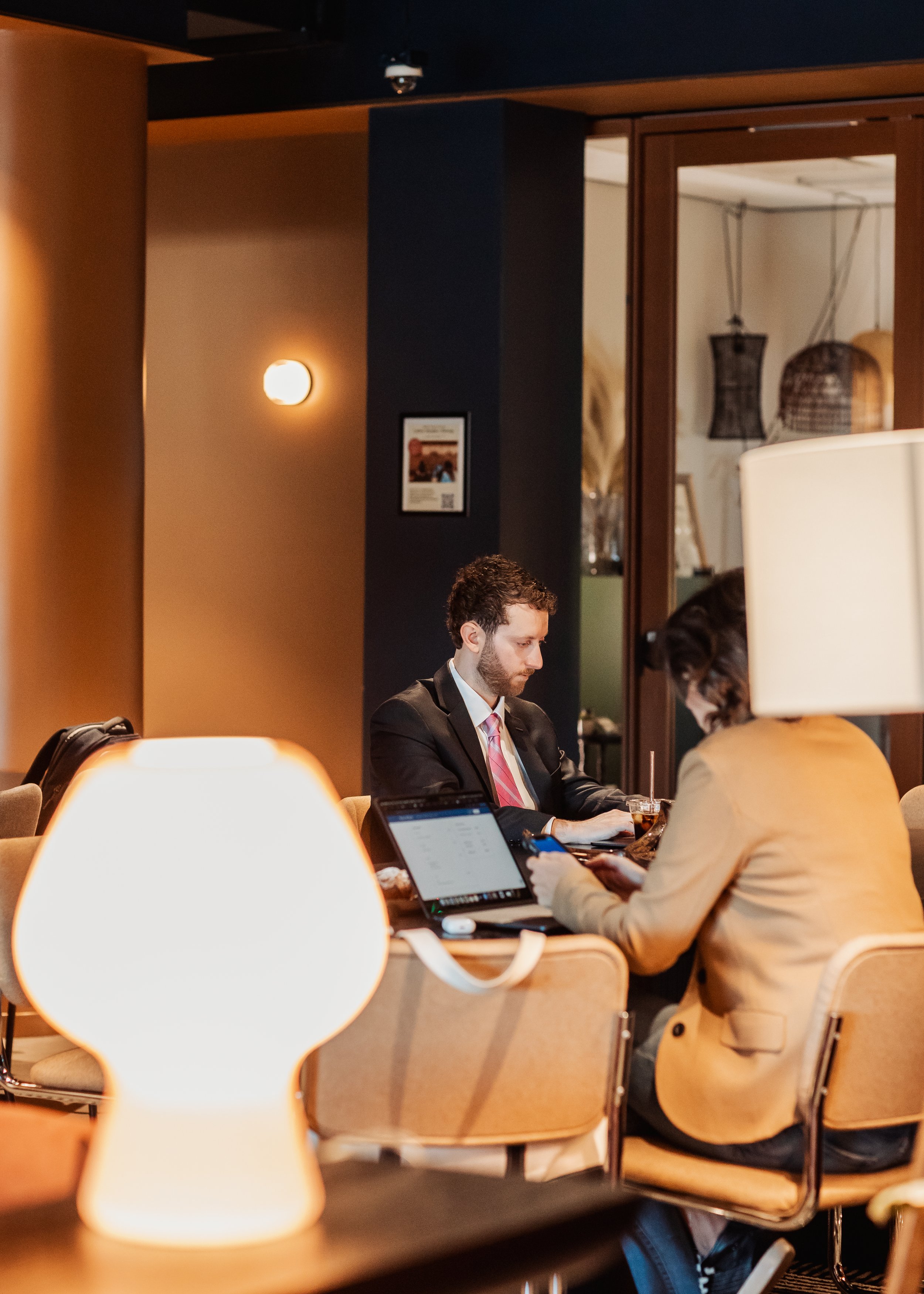 Two people sitting at a table in a restaurant or café, using electronic devices, with warm lighting and modern decor in the background.