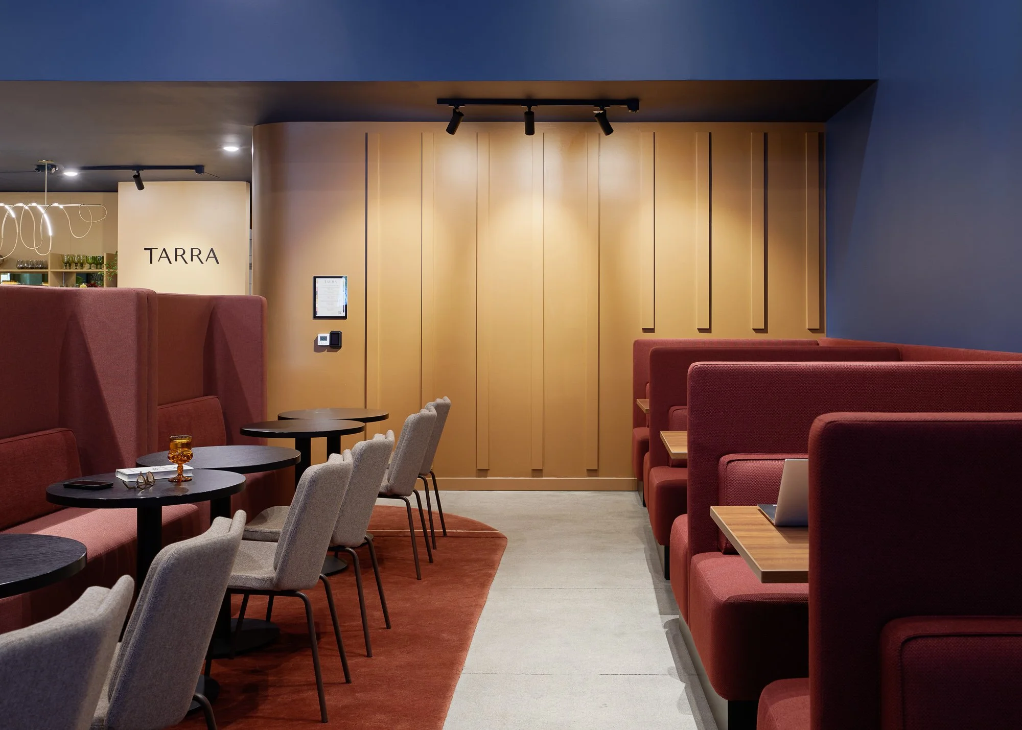Modern restaurant interior with beige and red seating, small black tables, and a sign that reads 'TARRA' on the wall.