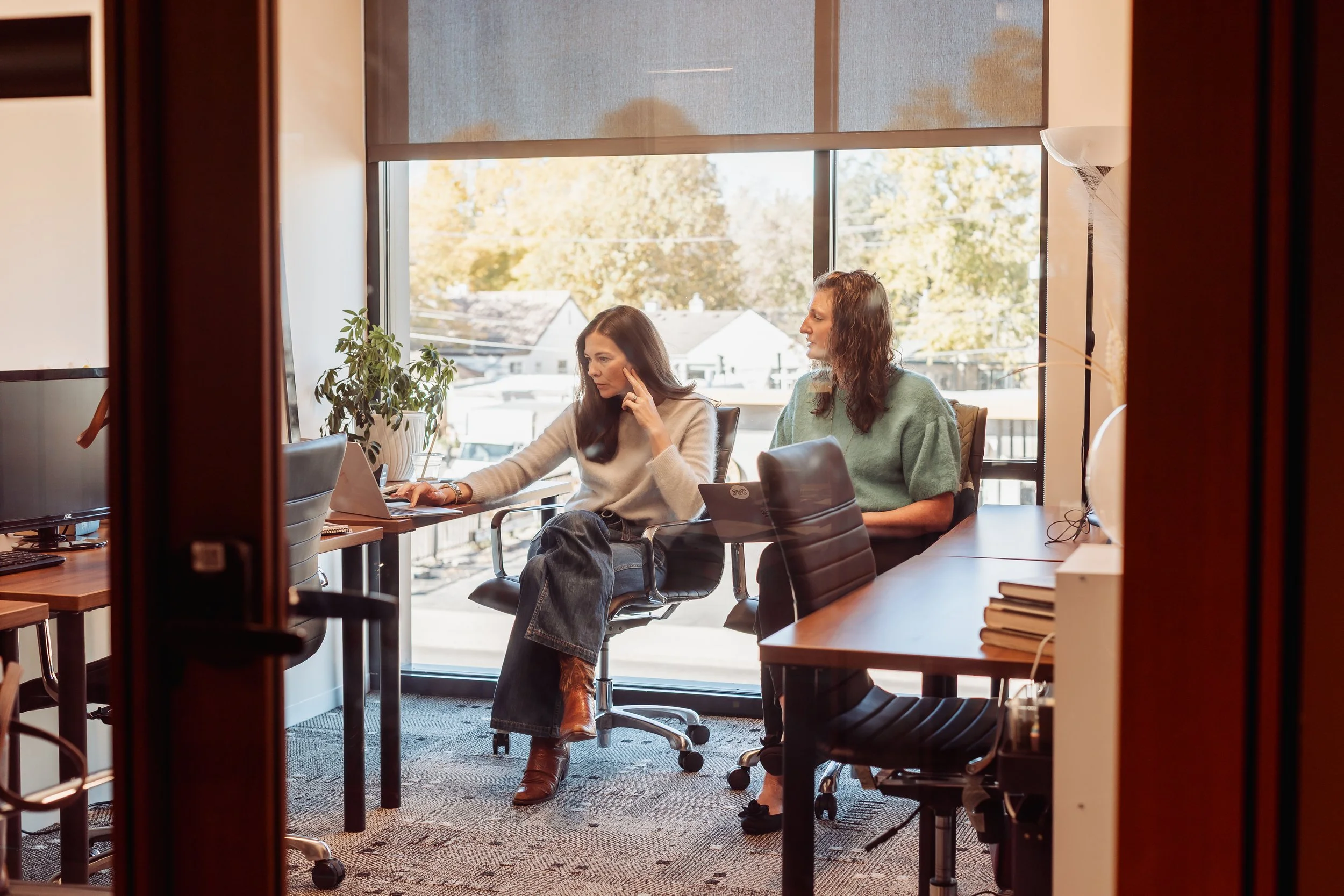Two women working in a modern office with large window showing outside trees and buildings. One woman is typing on a laptop, the other is looking at her screen, with a plant nearby.