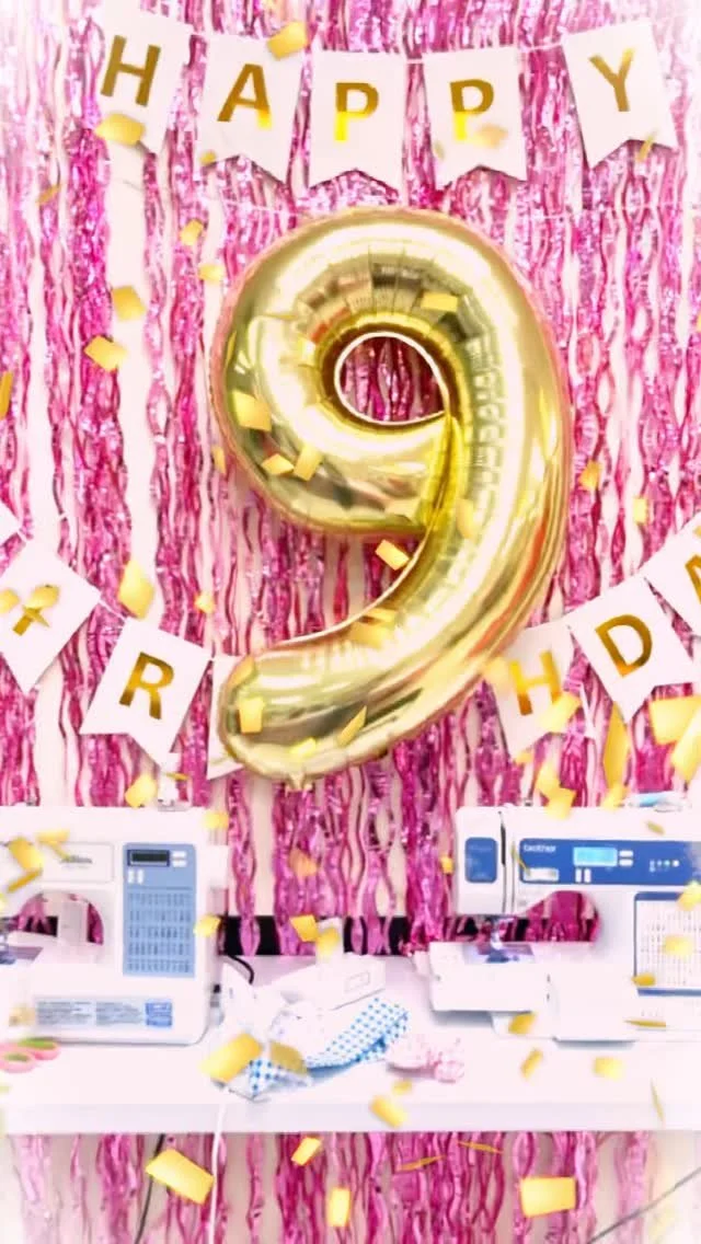 Decorative birthday celebration setup with a pink shiny backdrop, gold number nine balloon, gold letter banners spelling 'HAPPY BIRTHDAY', tissue paper confetti, and sewing machines on a table.