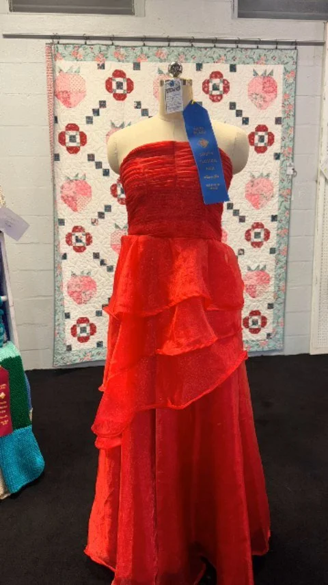 A red strapless gown with a layered skirt, displayed on a mannequin in front of a quilted wall hanging with a floral and heart pattern.
