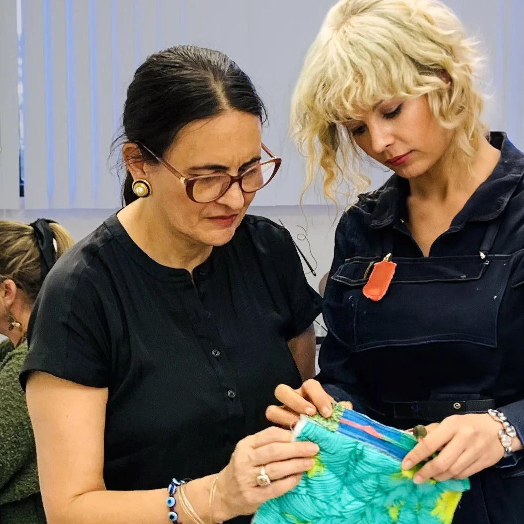 Two women examining a colorful fabric with a subdued background of vertical blinds.