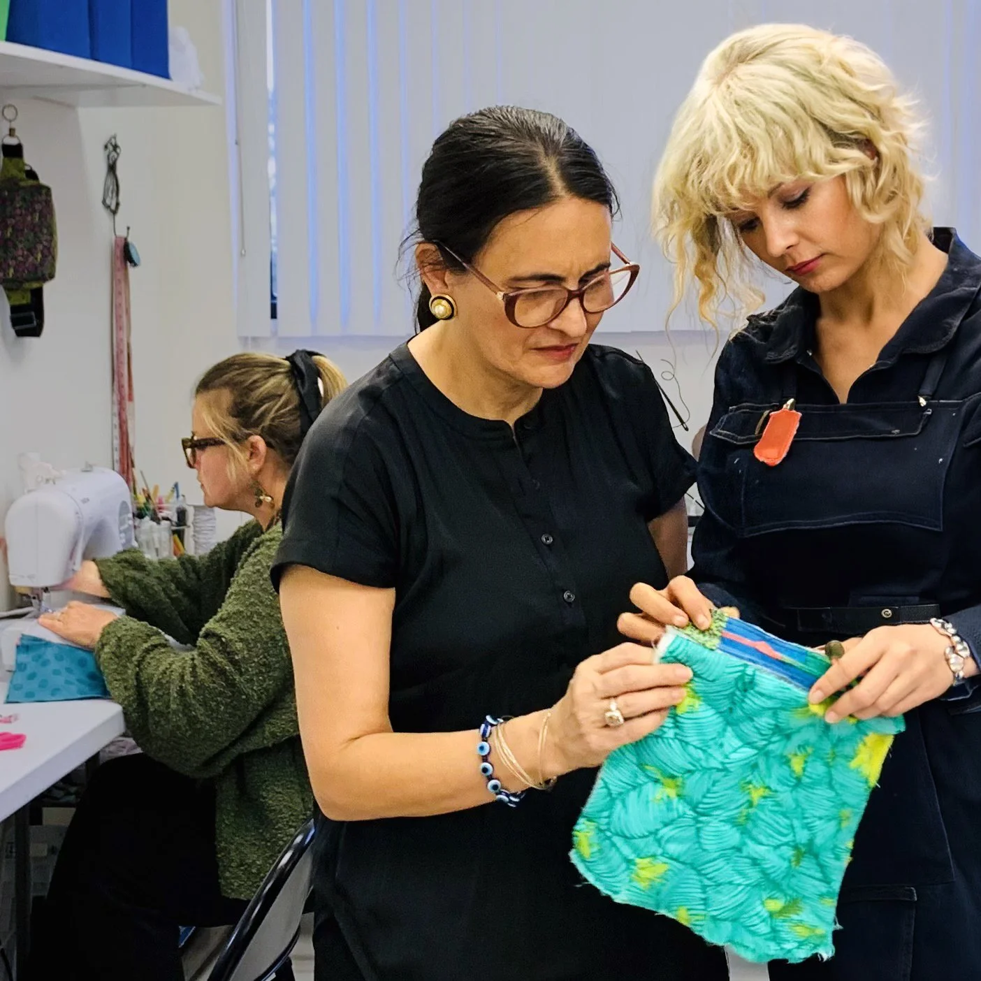 Two women working on a fabric project, one holding colorful fabric and the other assisting, with a woman sewing at a machine in the background.