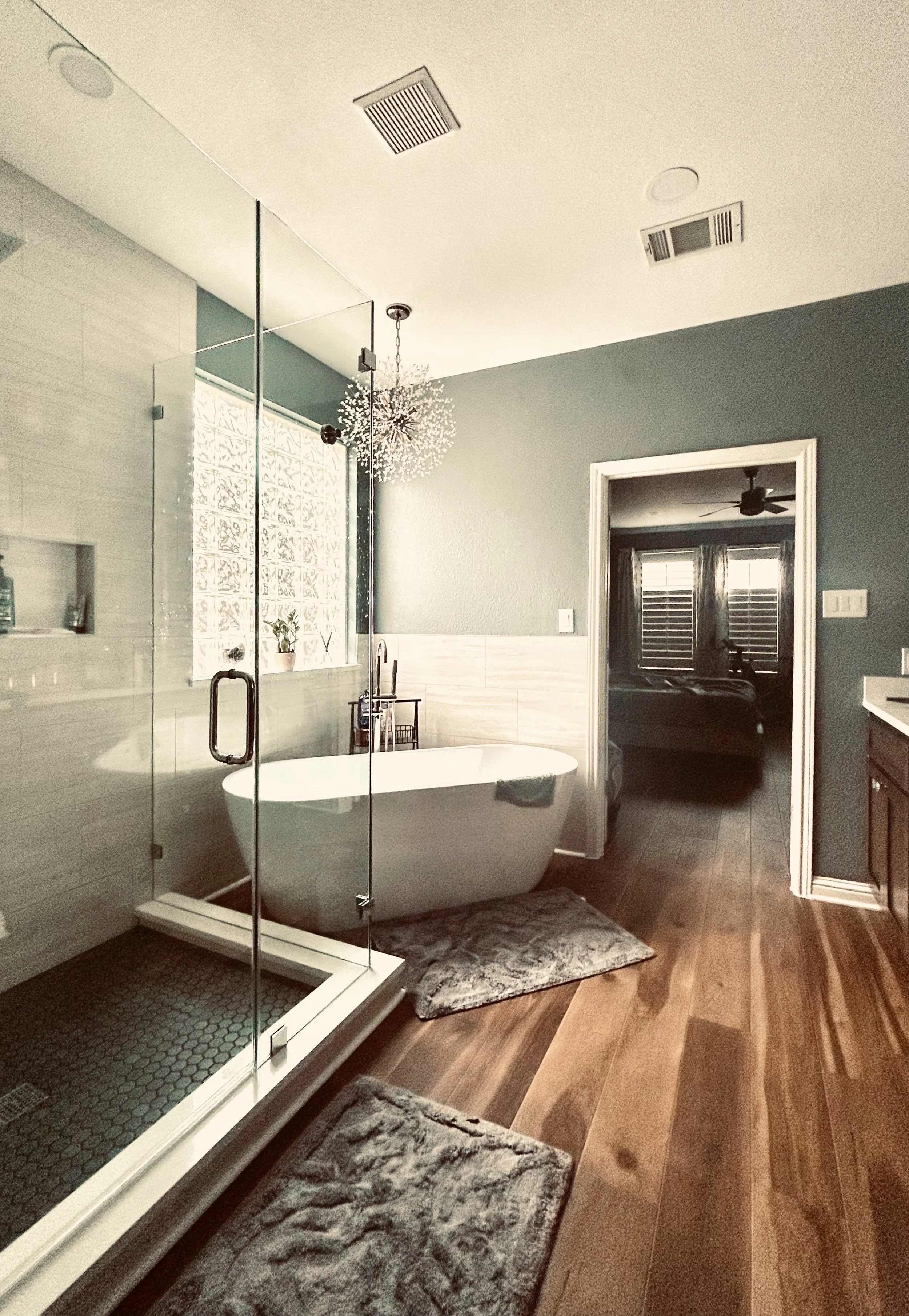 A modern bathroom with a freestanding bathtub, walk-in shower with glass door, wooden floor, and a blue wall with a chandelier hanging from the ceiling. There is a doorway leading to a room with shutters and a ceiling fan.