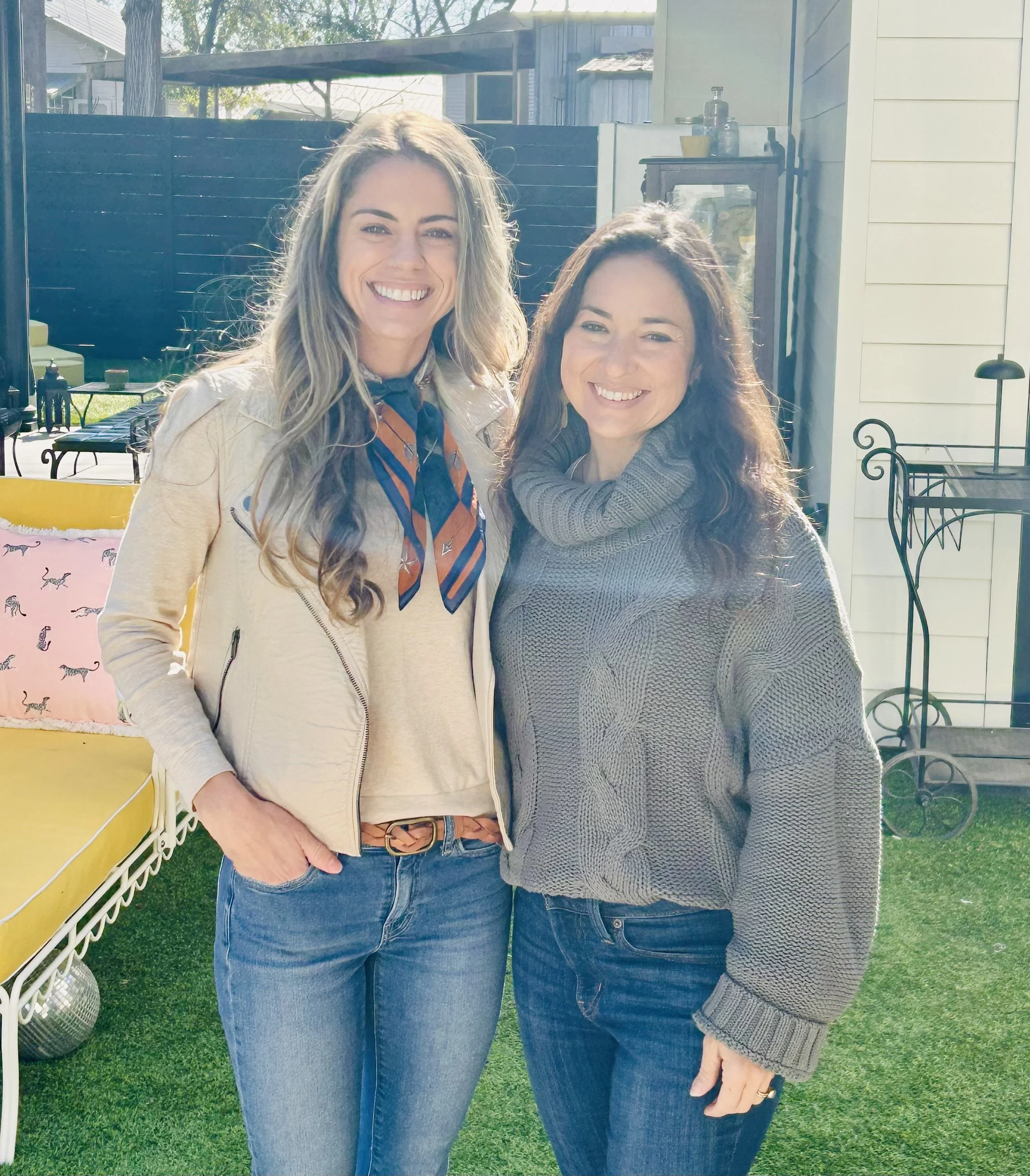 Two women smiling outdoors on a sunny day, standing close together in a backyard with a yellow bench and green grass.
