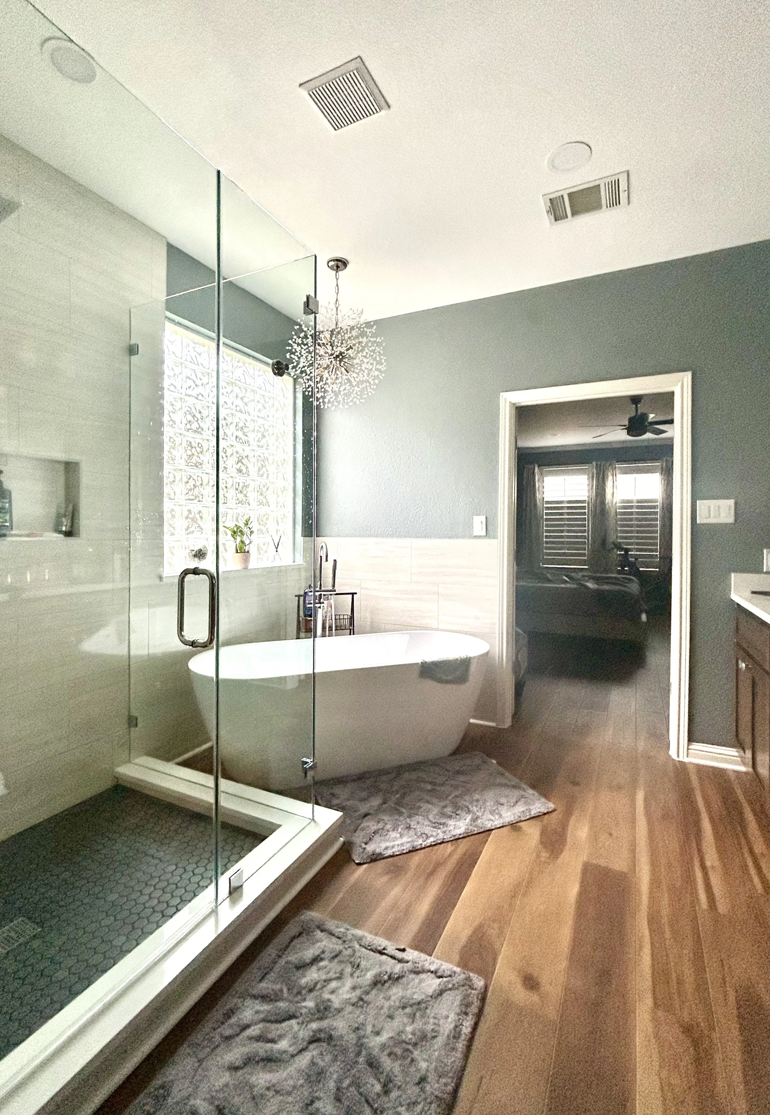 Modern bathroom with a glass-enclosed shower, a freestanding bathtub, and a wooden floor. The bathroom has a chandelier, a window with glass blocks, and an open door leading to a bedroom with two windows and a ceiling fan.