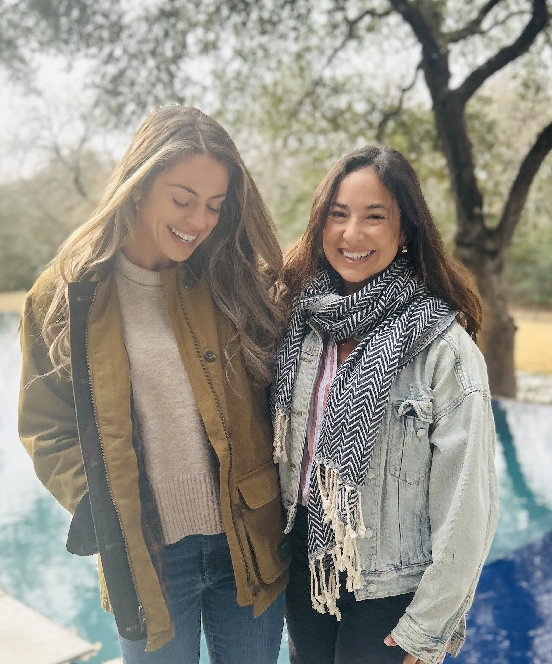 Two women smiling and standing close together outdoors near water, with trees in the background.