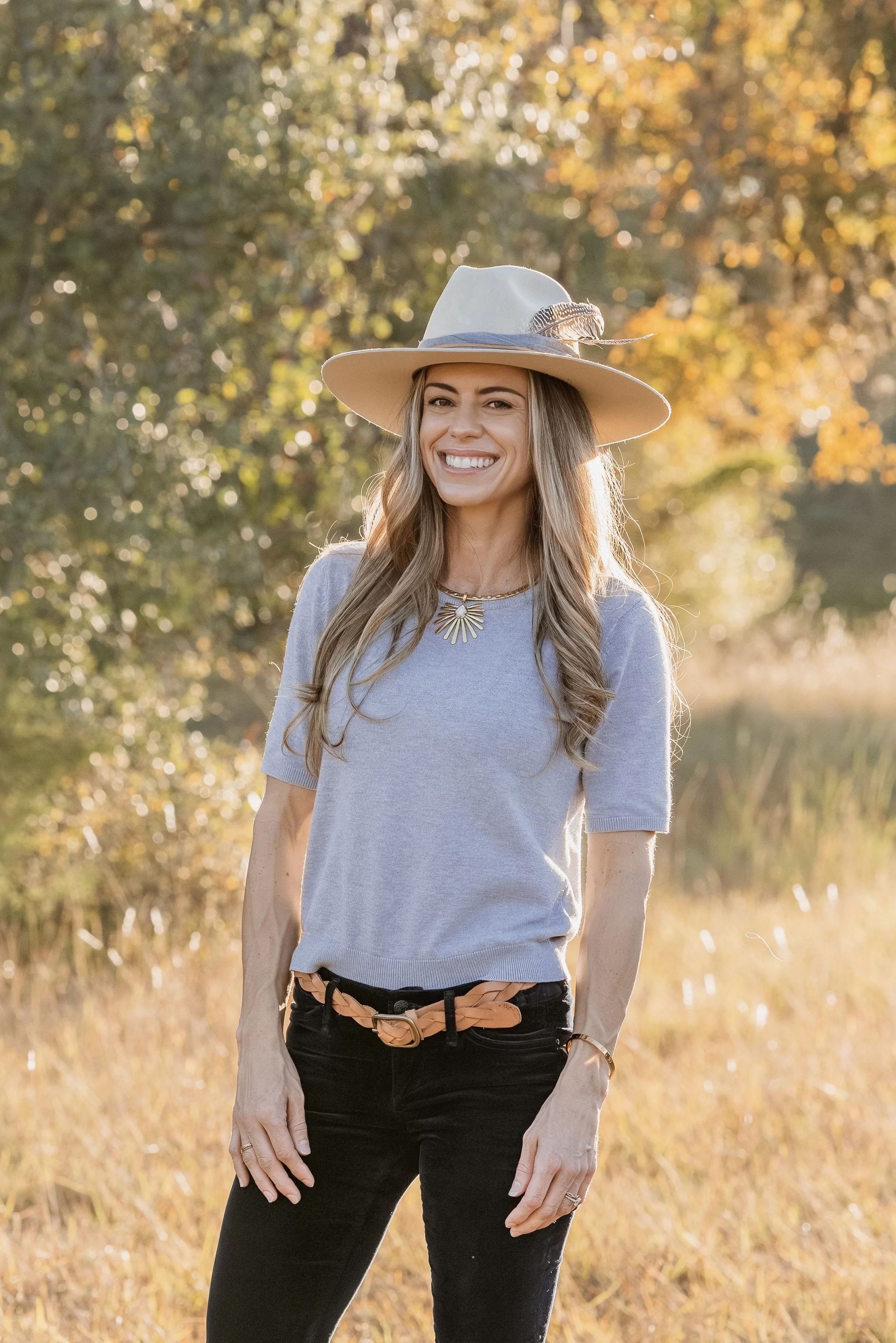 A woman wearing a wide-brimmed hat with a feather, a light gray short-sleeved sweater, black jeans with a belt, and a necklace, standing outdoors in a sunny, autumn landscape with trees and grass.