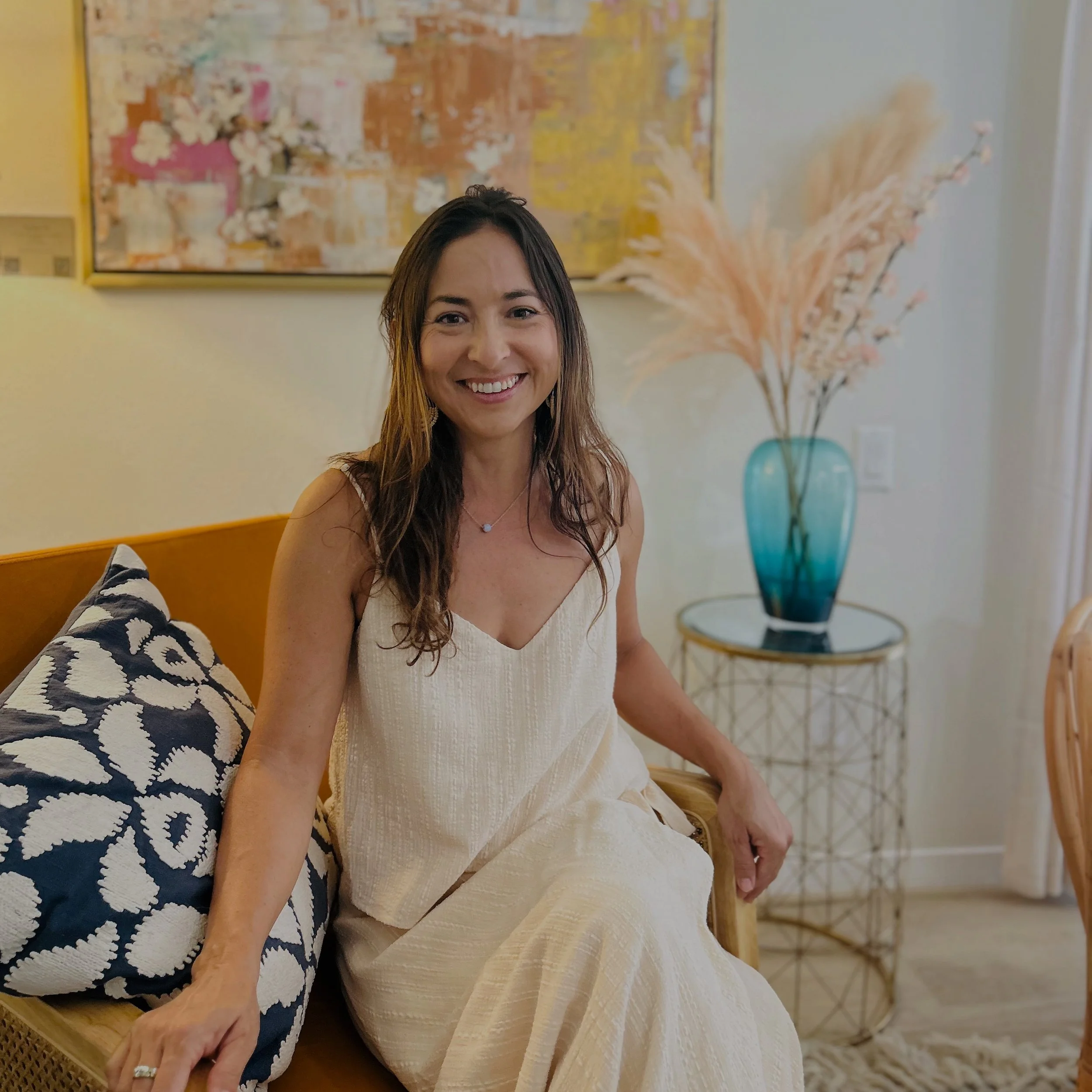 A woman with long brown hair sitting on a beige sofa, smiling at the camera, in a room decorated with a colorful abstract painting, a blue vase with pampas grass, and a side table.