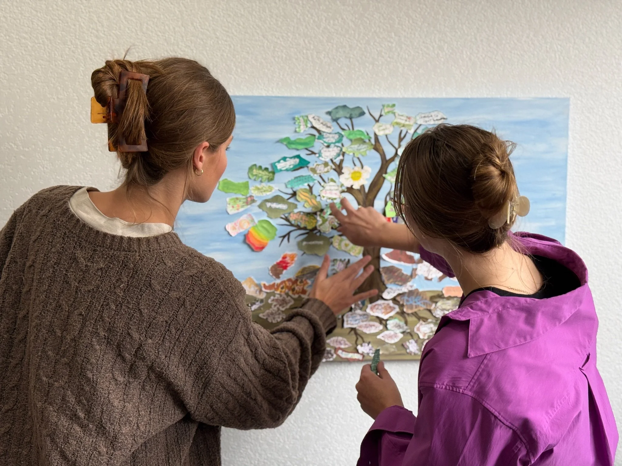 Two women are looking at a colorful art piece of a tree on a wall. One woman is wearing a brown sweater and the other is wearing a purple jacket.