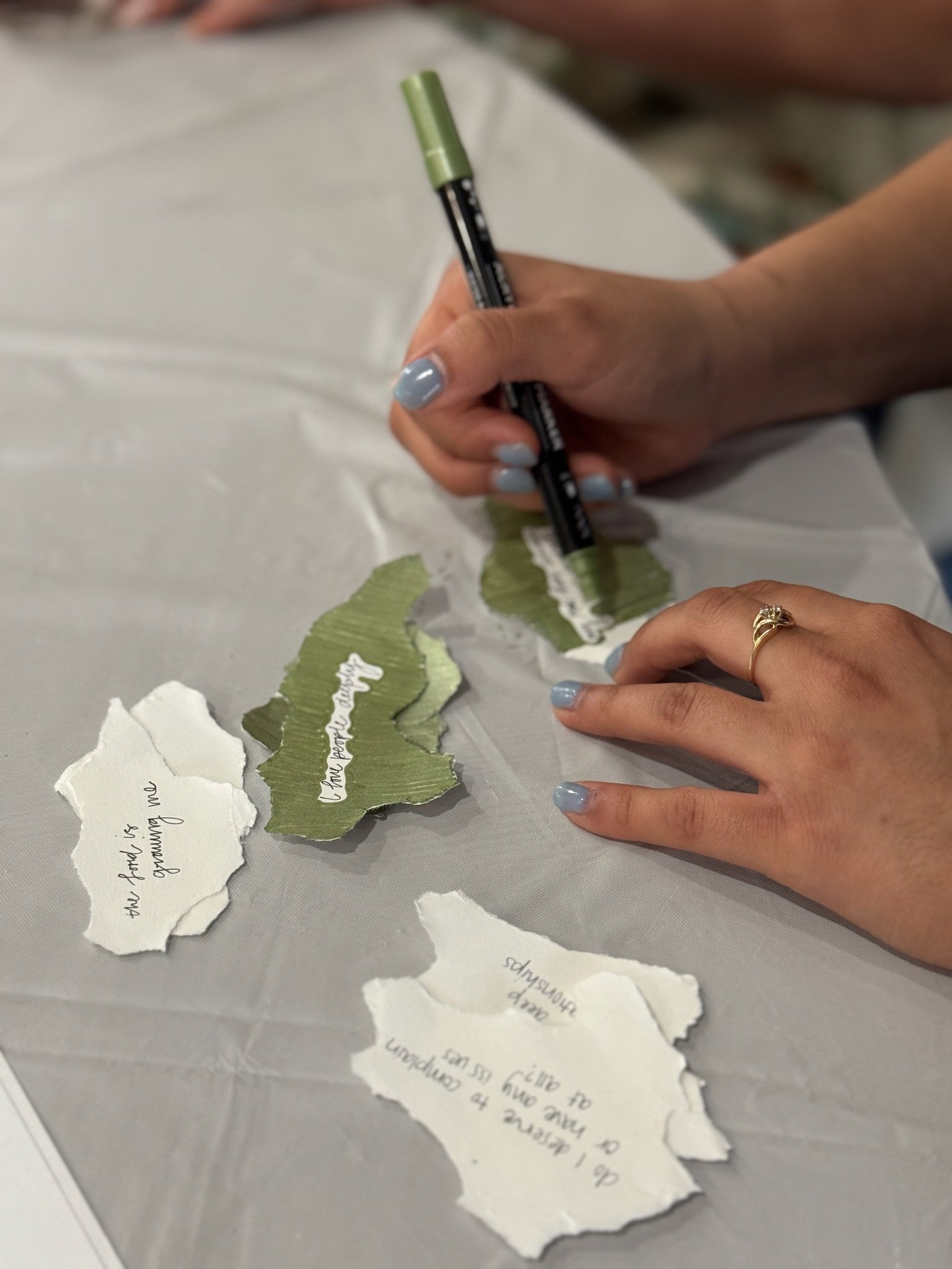 A person with painted nails wearing a gold ring is holding torn pieces of paper with handwritten notes in black ink. The torn papers are light green and white, with some text visible.
