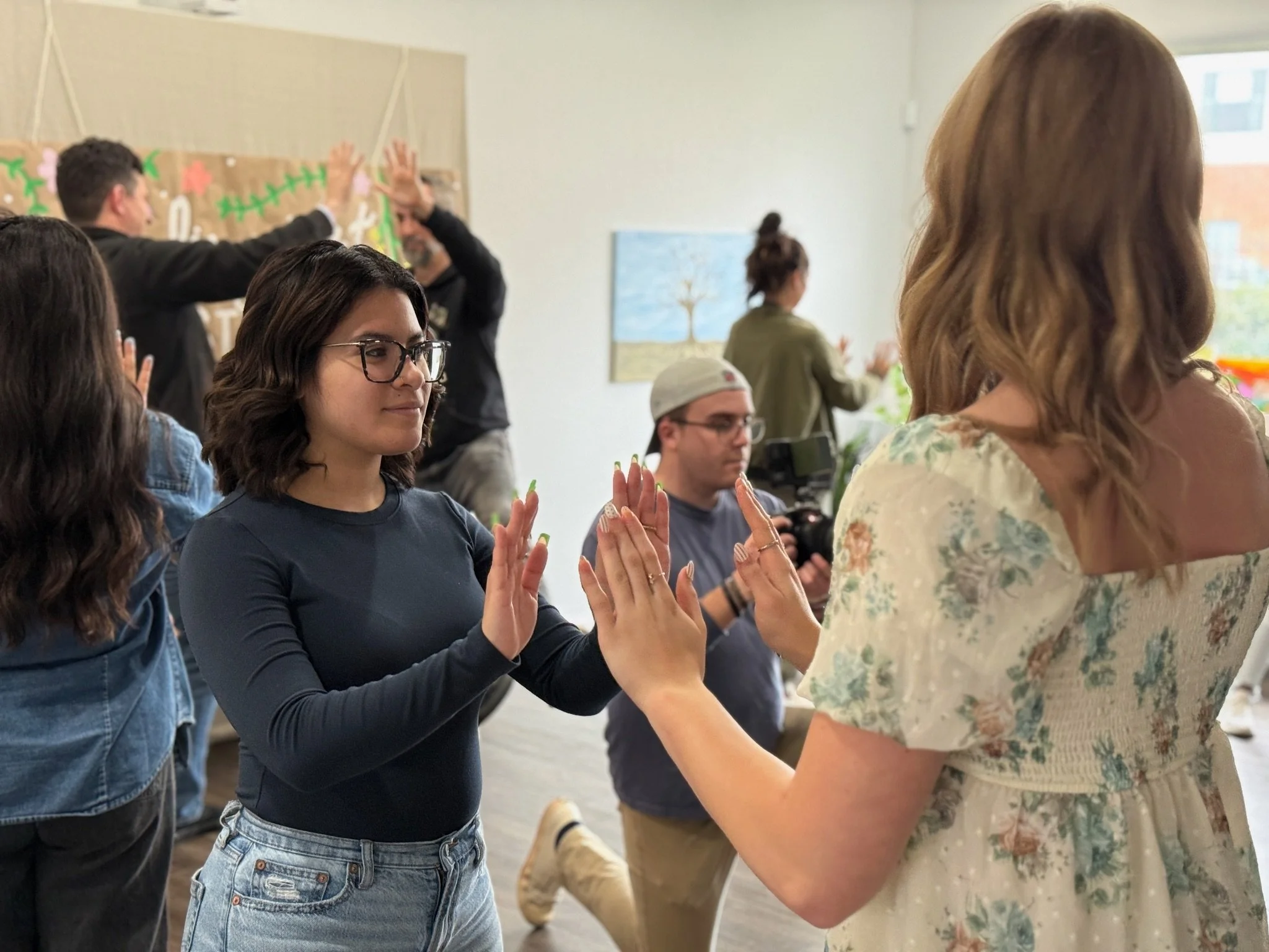 People participating in a group activity, holding hands, in a well-lit room with a colorful wall decoration and a painting of a tree on the wall.
