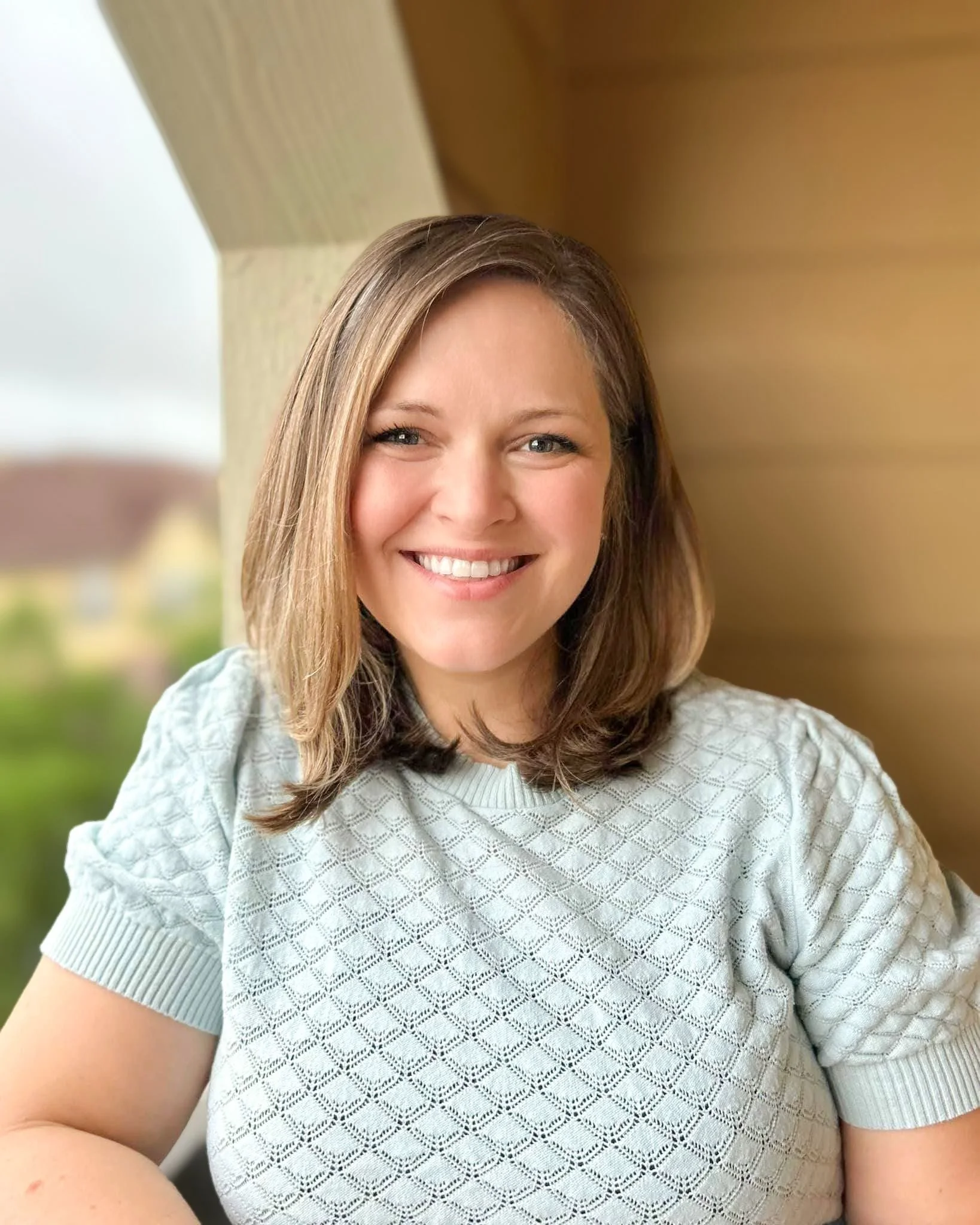 Close-up of smiling woman with light brown hair wearing a white textured short-sleeve top, outdoors with blurred background.
