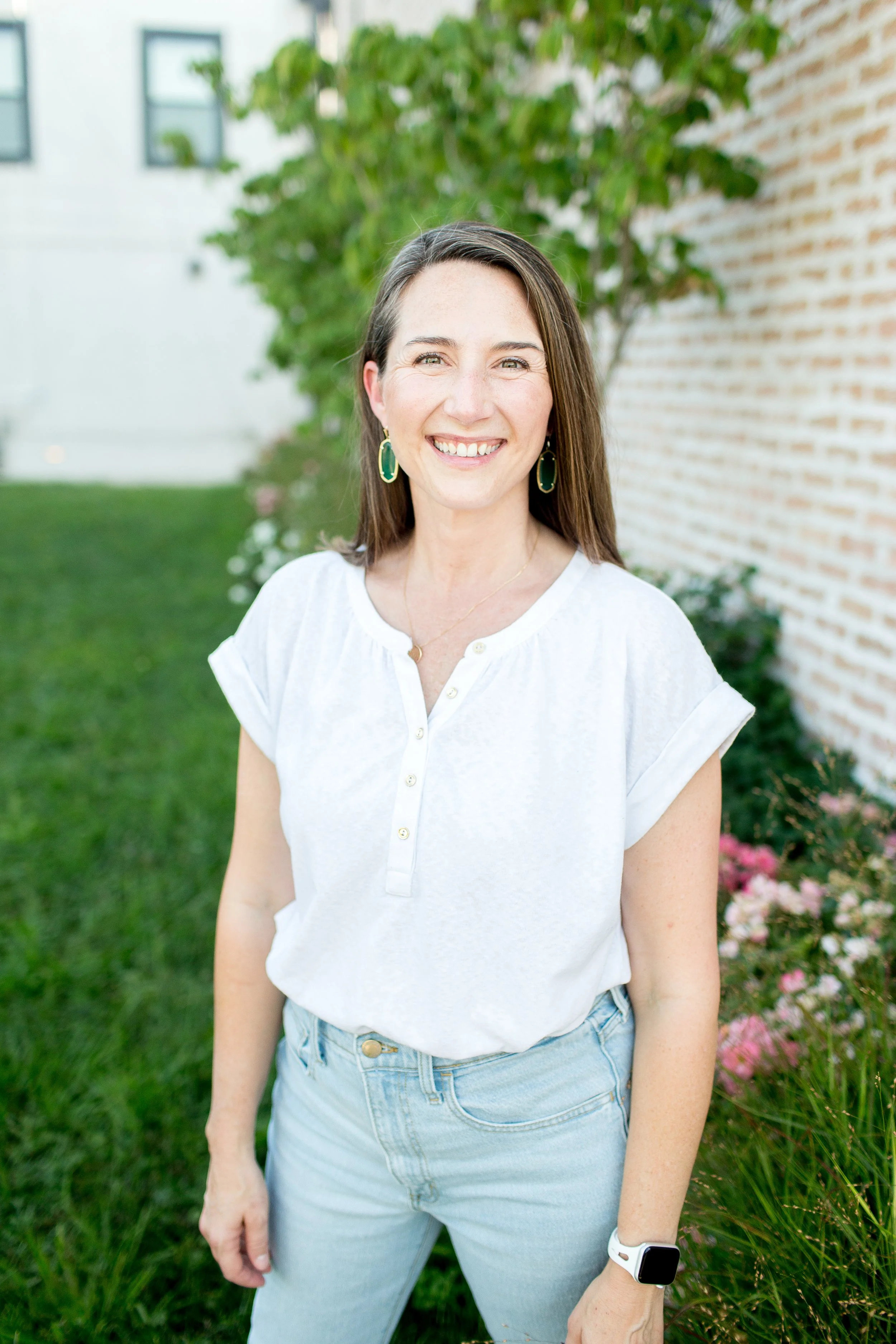 A woman with long brown hair smiling outdoors, wearing a white button-up shirt, light blue jeans, green earrings, and a smartwatch, standing on grass with a brick wall and green plants in the background.