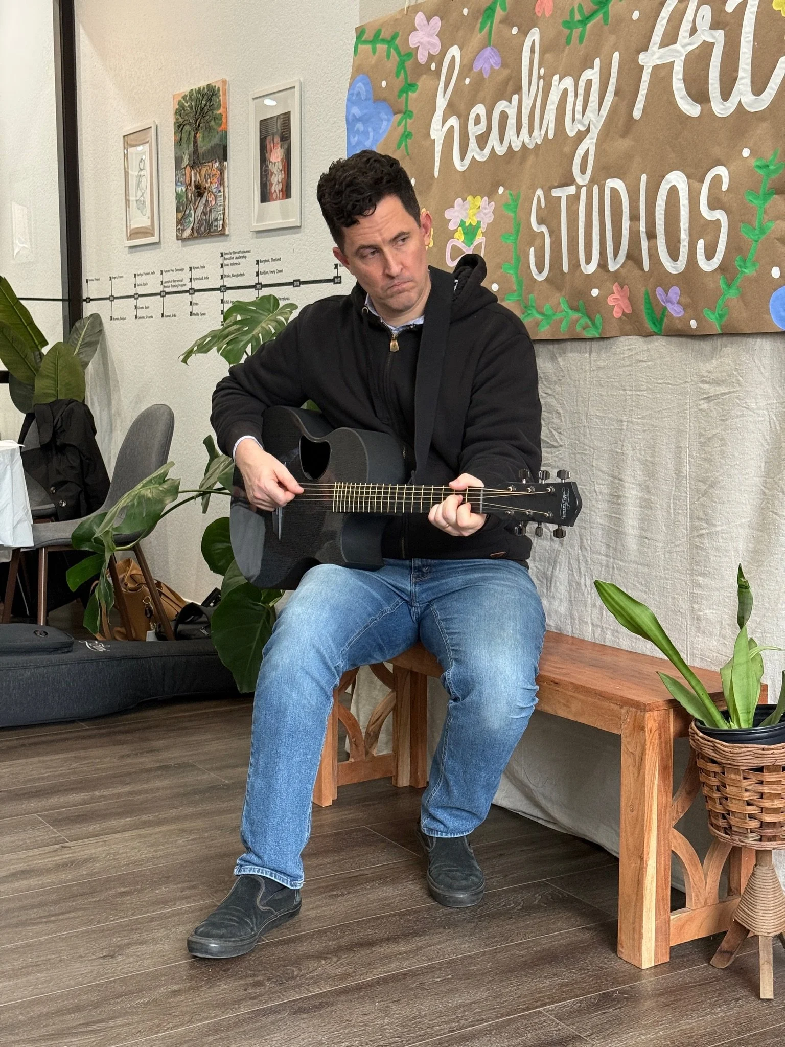 Man sitting on a wooden bench playing an acoustic guitar in a room with plants and colorful wall art.