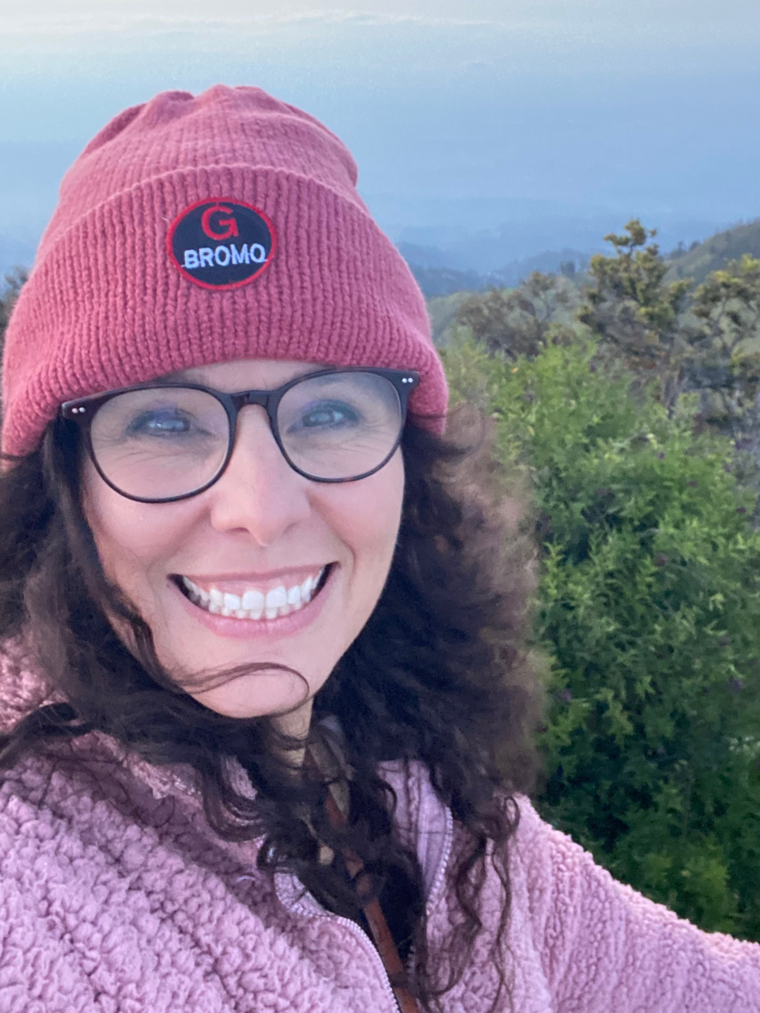 Close-up of a woman smiling outdoors wearing glasses and a pink knit beanie with a patch that says 'G Bromo', and a pink fleece jacket, with trees and mountains in the background.