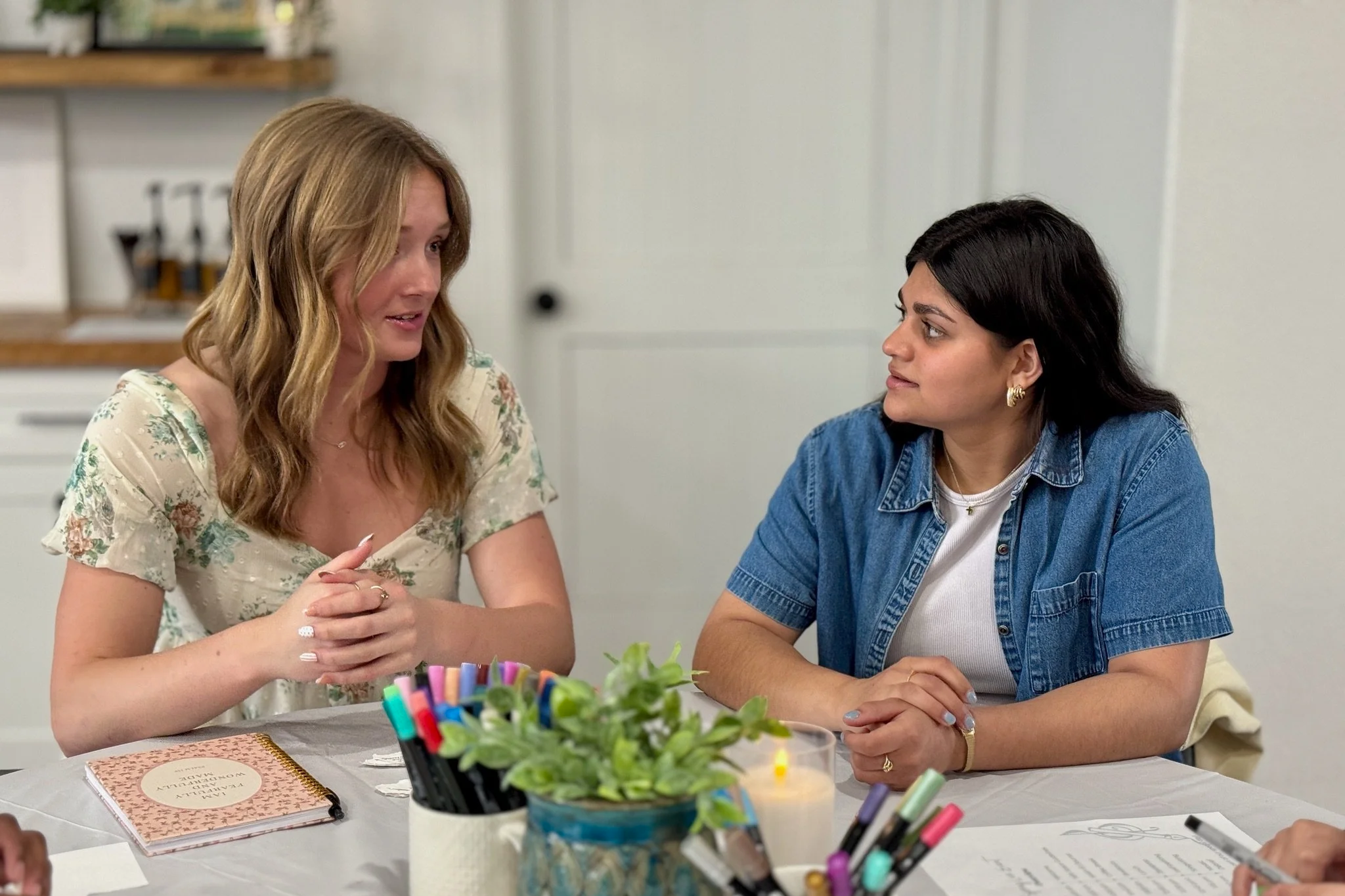 Two women sitting at a table engaged in conversation. One woman has long, wavy blonde hair and is wearing a floral patterned top; the other has shoulder-length dark hair and is wearing a denim jacket. The table has colorful markers, a notebook, and a plant in the foreground.