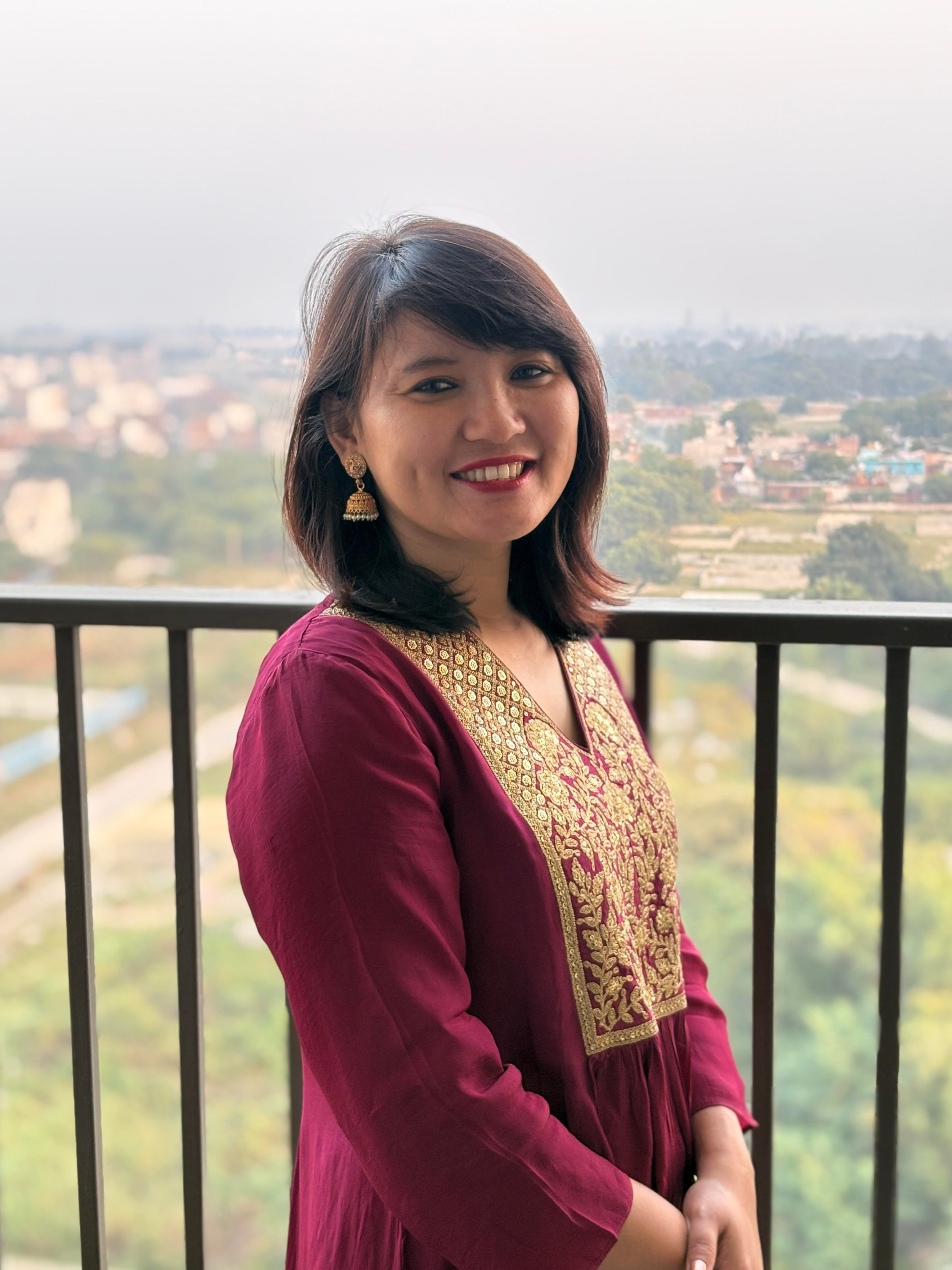 Woman in traditional South Asian attire standing outdoors with a cityscape and greenery in the background.