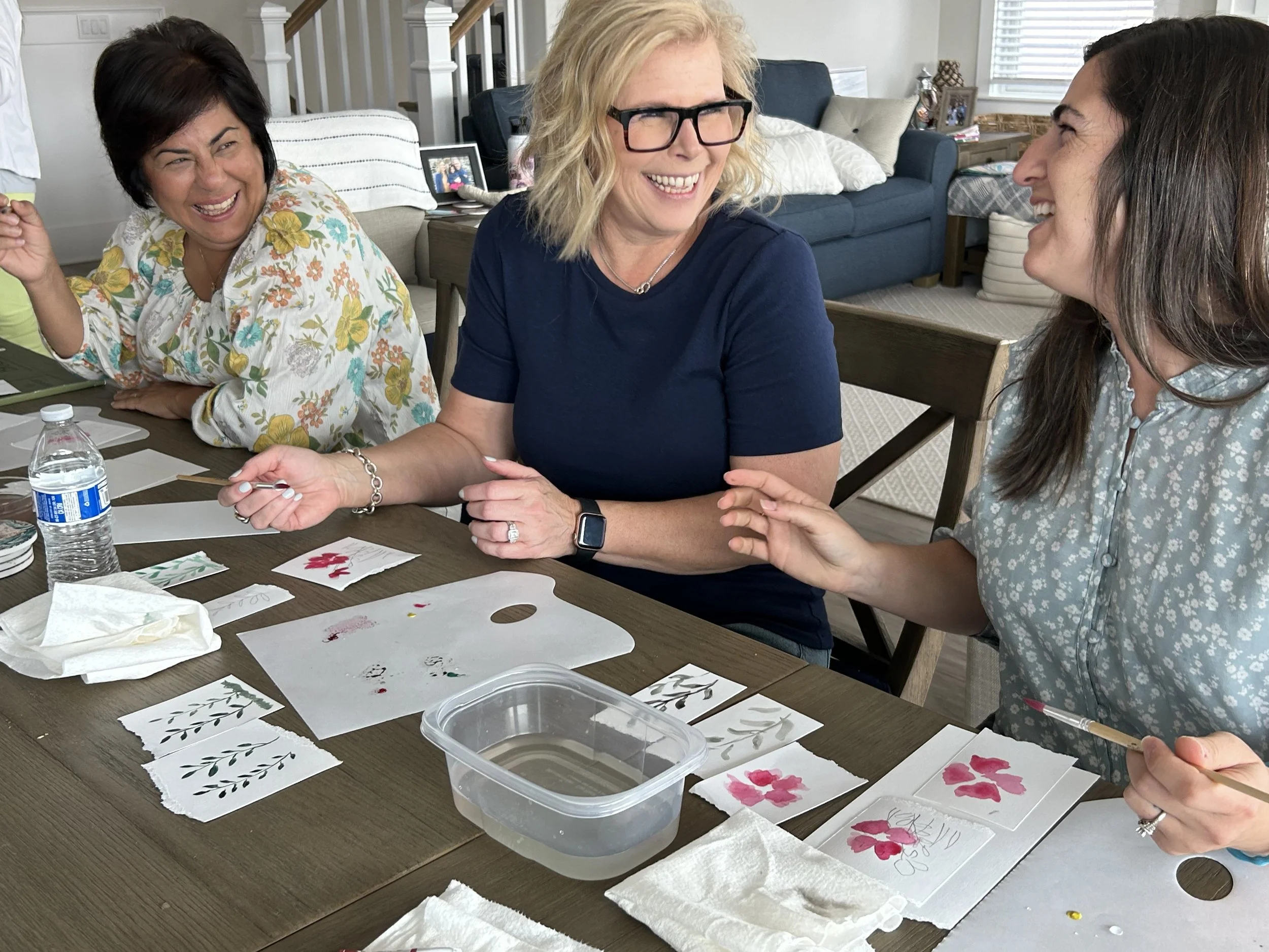 Three women are sitting at a dining table engaging in arts and crafts, with watercolor paintings of flowers and leaves in front of them. They are smiling and laughing, enjoying their creative activity.