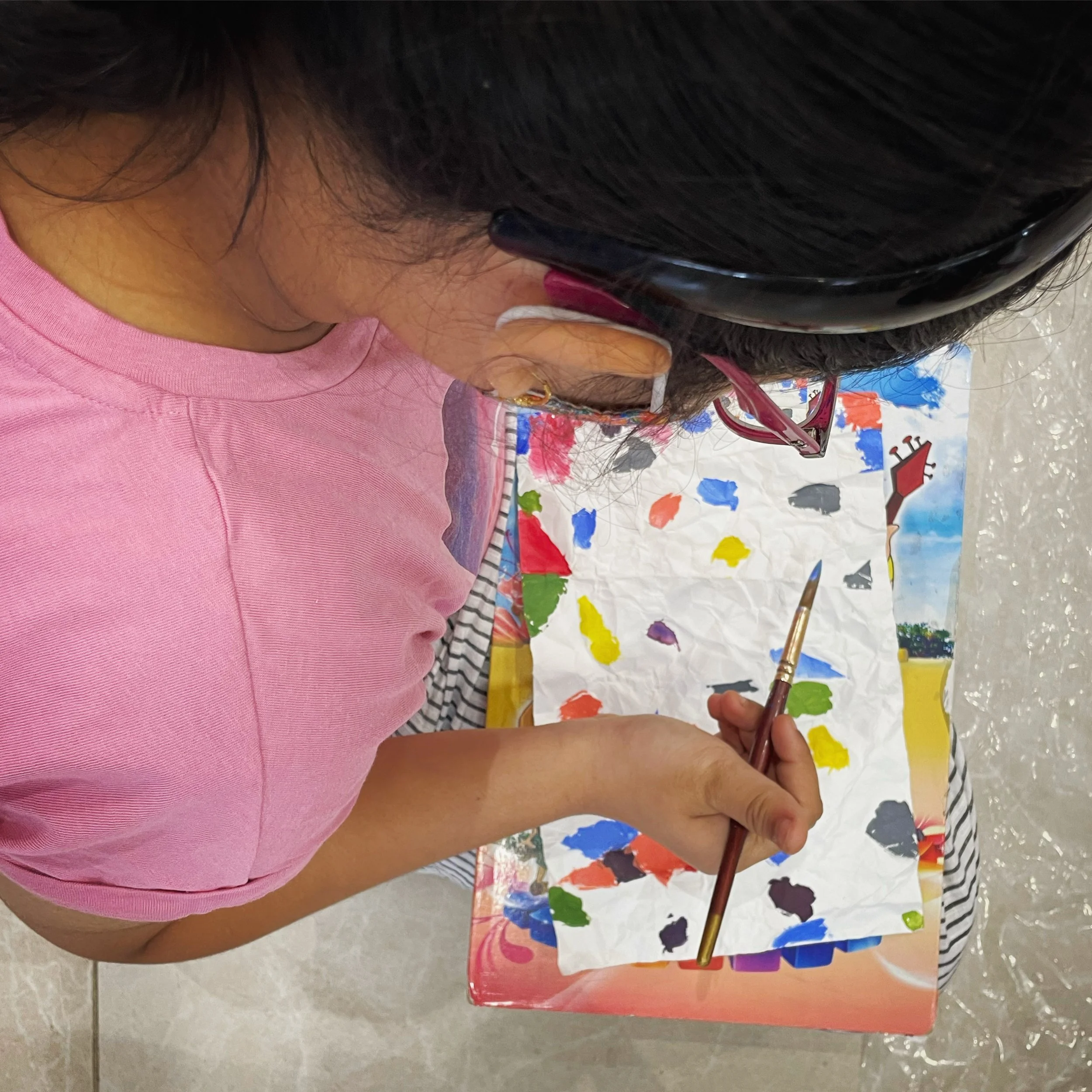 A person with long dark hair wearing a pink top and striped shirt, painting on a colorful palette with various paint colors and a paintbrush in hand.