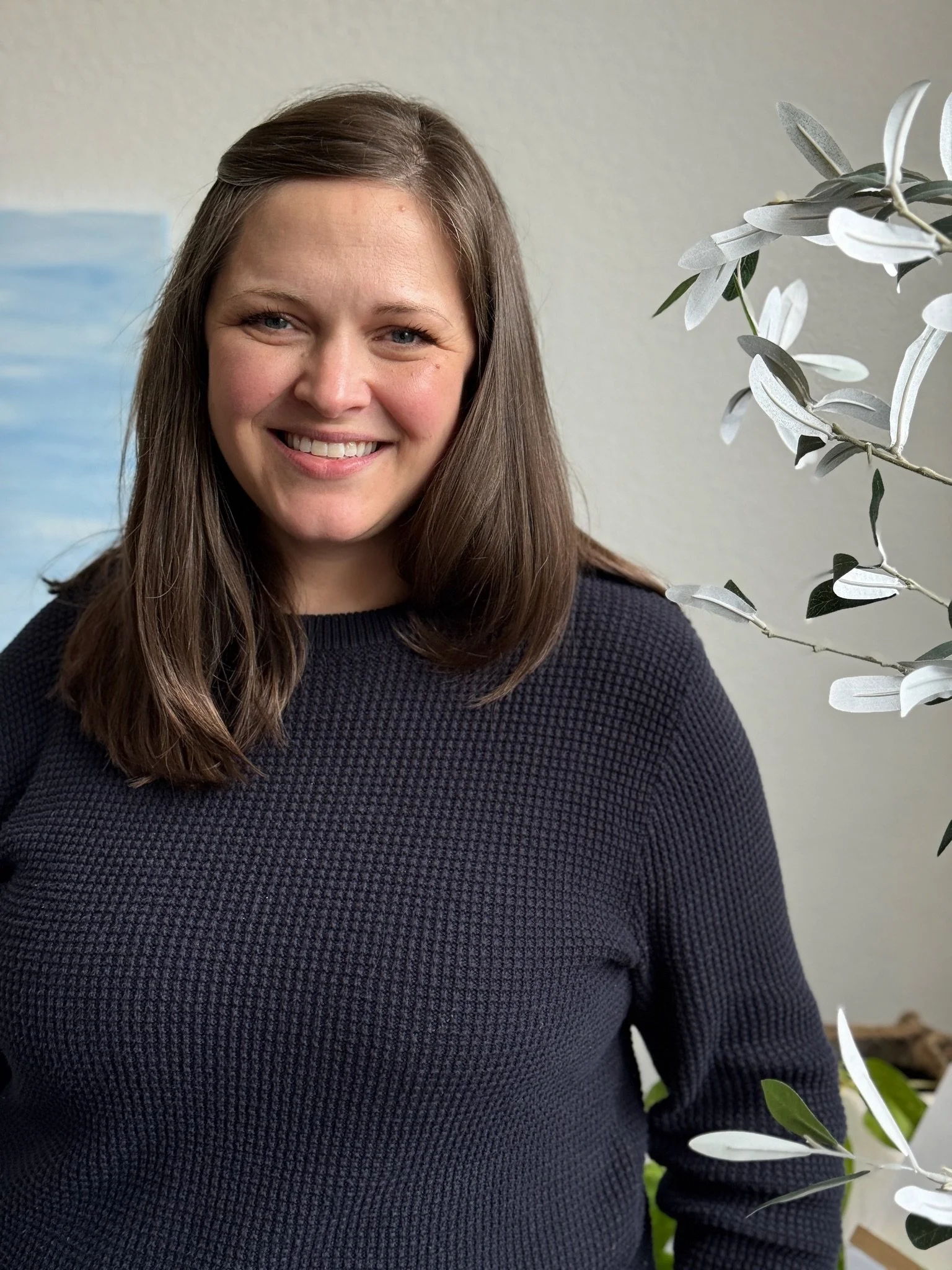 A smiling woman with long brown hair in a black sweater standing indoors next to a houseplant with silvery-green leaves.