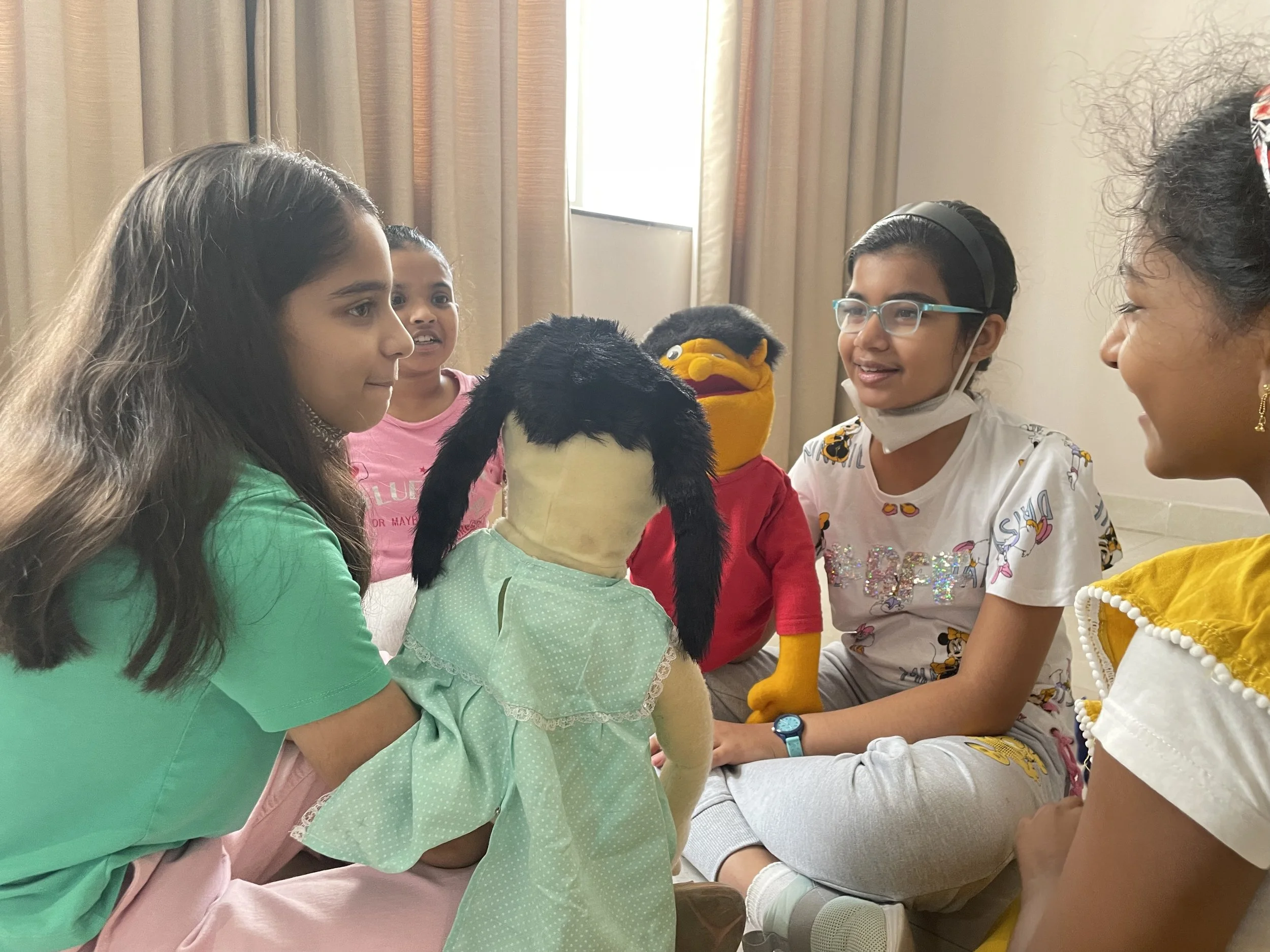 Children sitting in a circle, playing with puppets in a room with curtains.