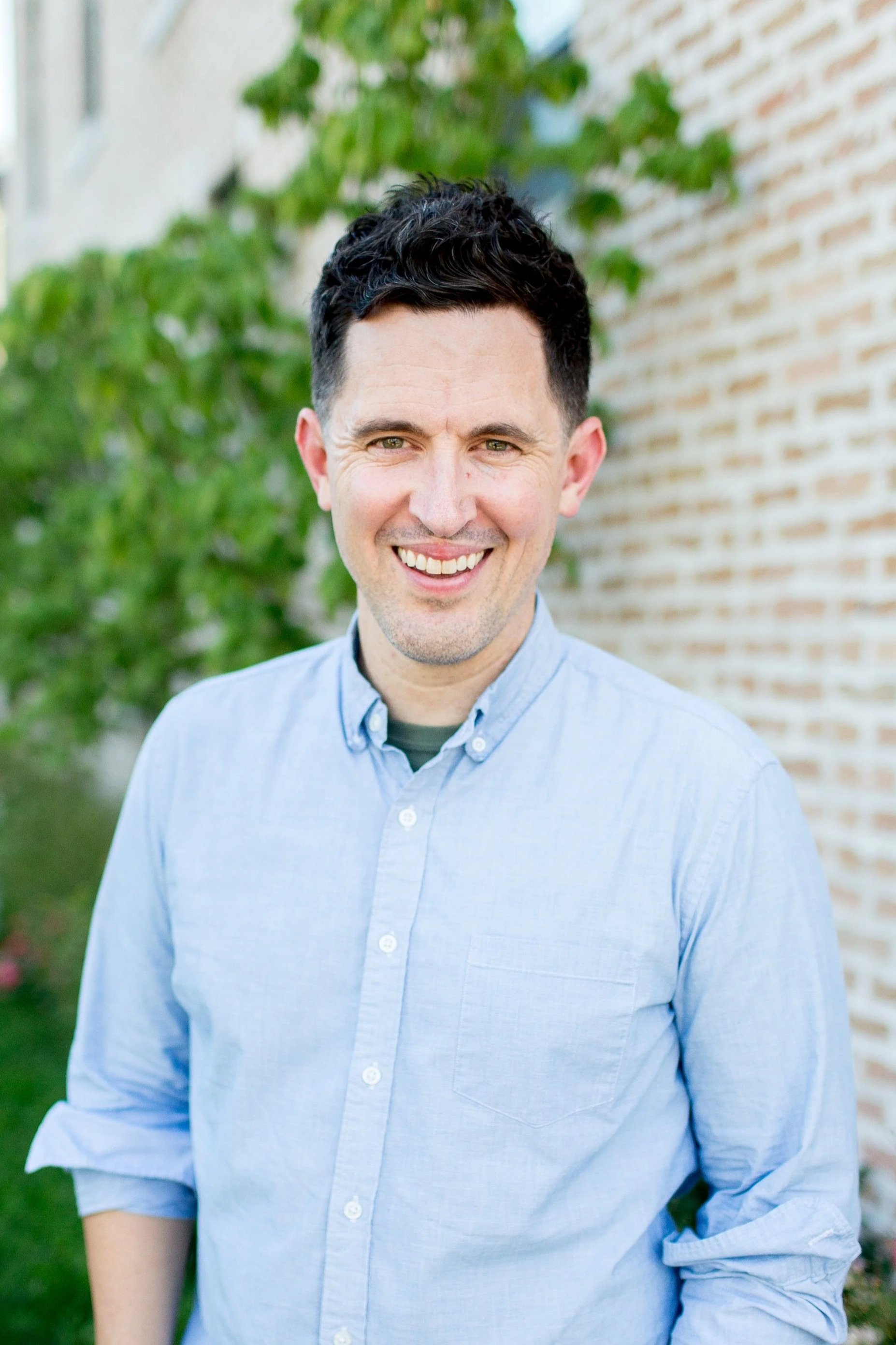 Portrait of a young man with dark, curly hair and green eyes smiling outdoors. He is wearing a light blue button-up shirt, with a brick wall and green foliage in the background.