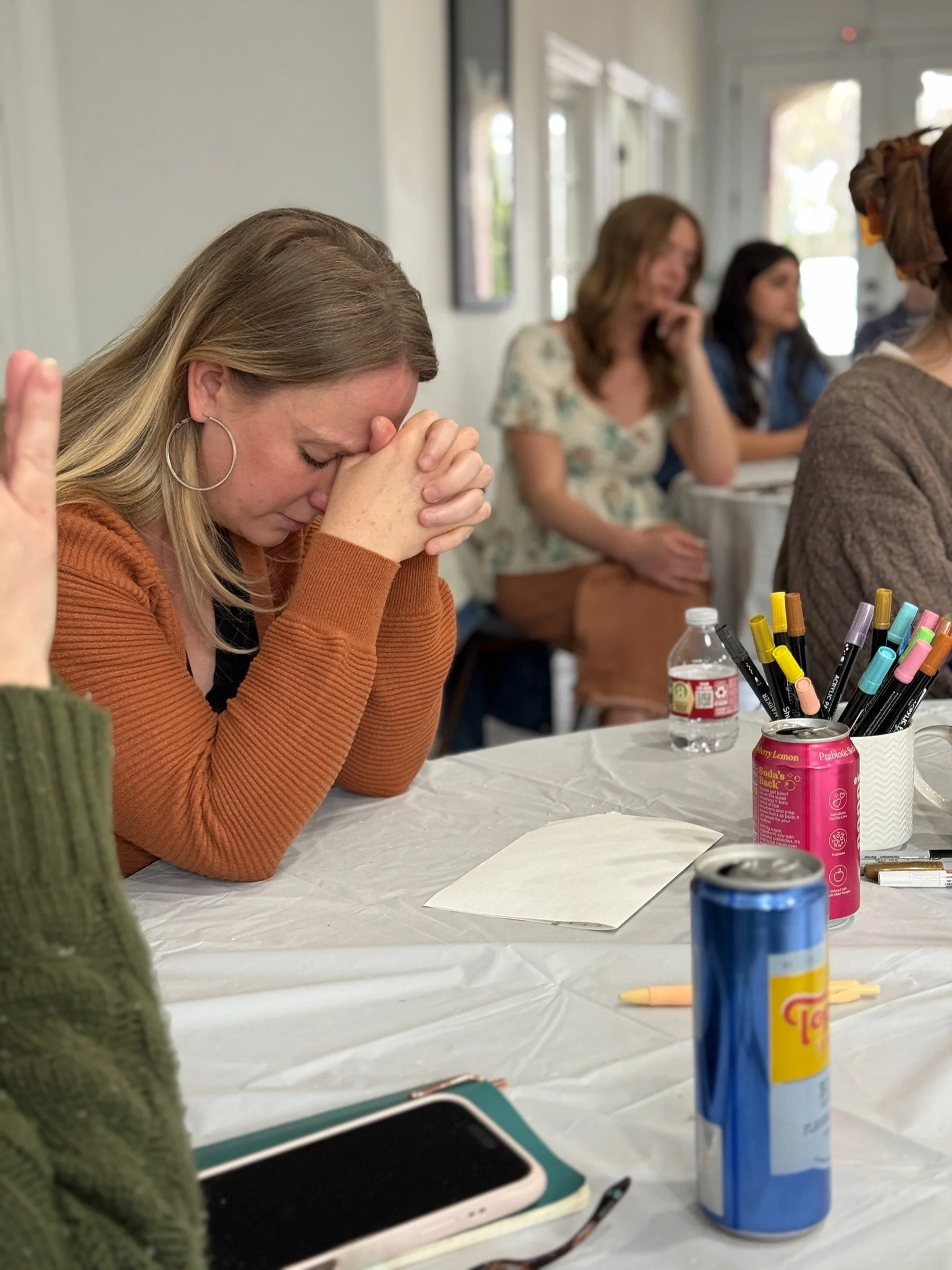 A woman with long blonde hair and hoop earrings is sitting at a table with her forehead resting on her clasped hands, appearing emotional or thoughtful, surrounded by art supplies in a bright room.
