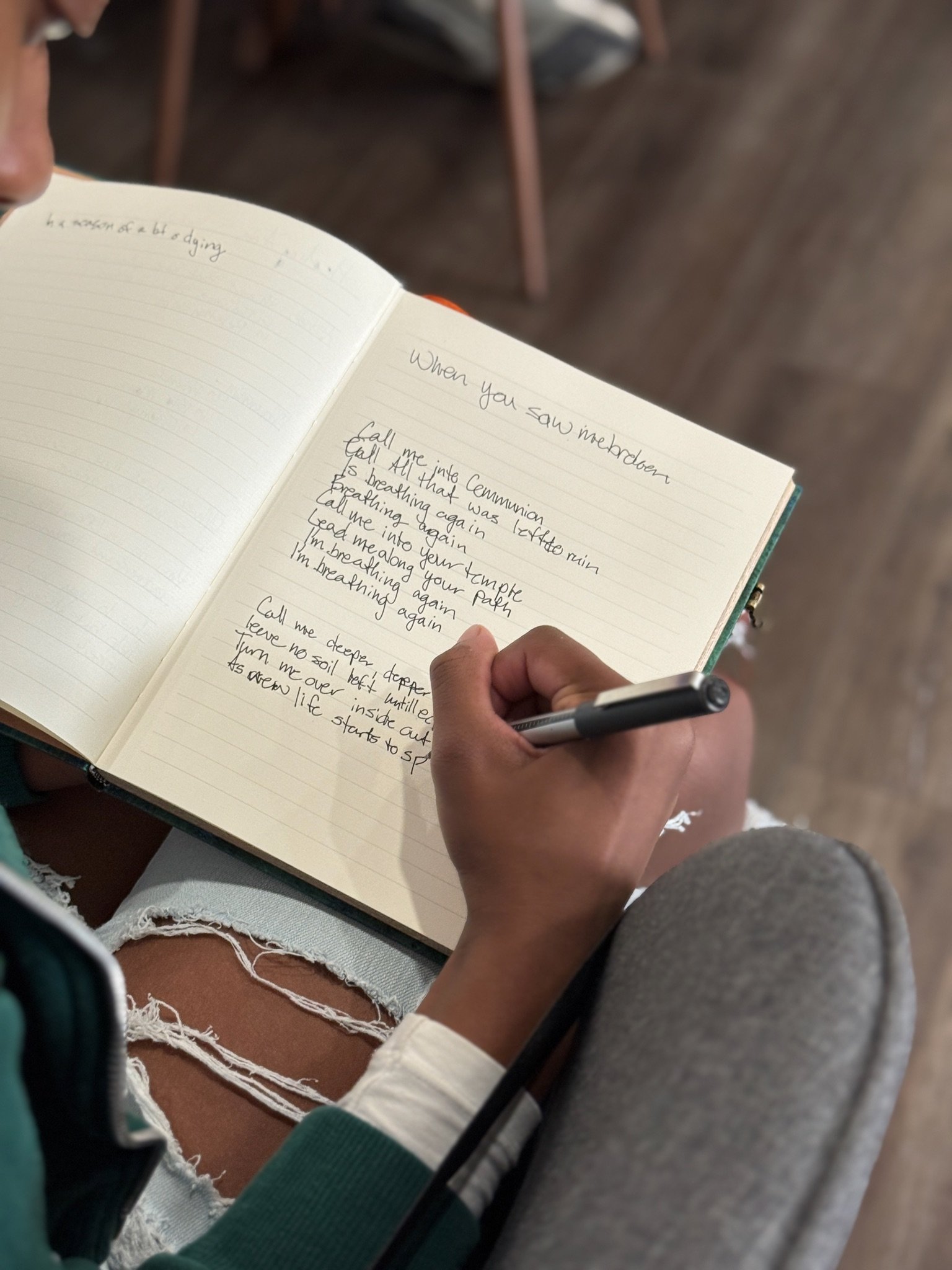 A person sitting on a chair, holding a pen and writing in a notebook with handwritten text, wearing ripped jeans and a green long-sleeve shirt, with other chairs and a wooden floor in the background.