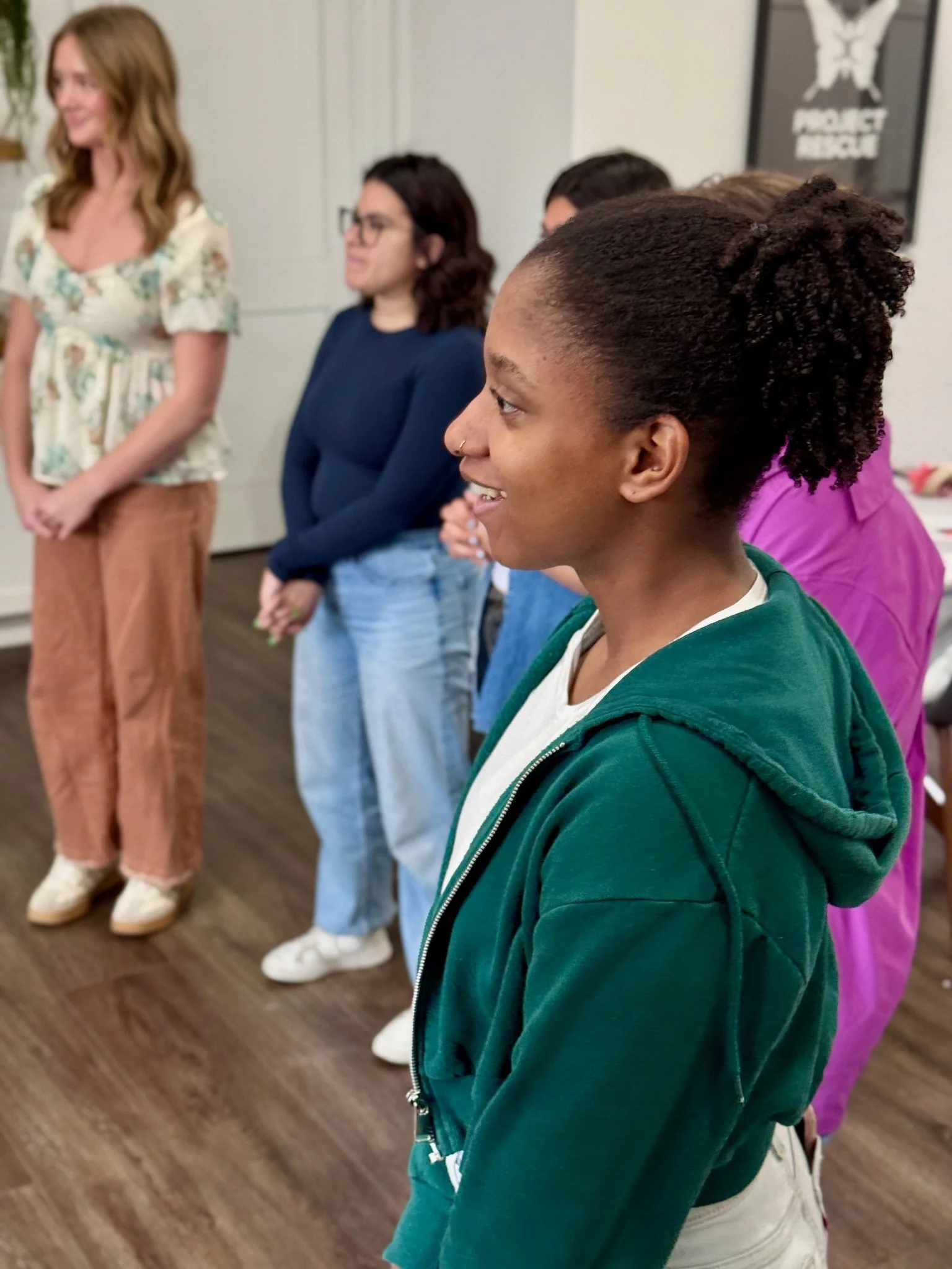 A group of people standing indoors, with a woman in a floral shirt and tan pants standing in the background, and a woman with dark hair, glasses, wearing a navy shirt. In the foreground, a woman with natural curly hair styled in a puff, wearing a green hoodie and white pants.