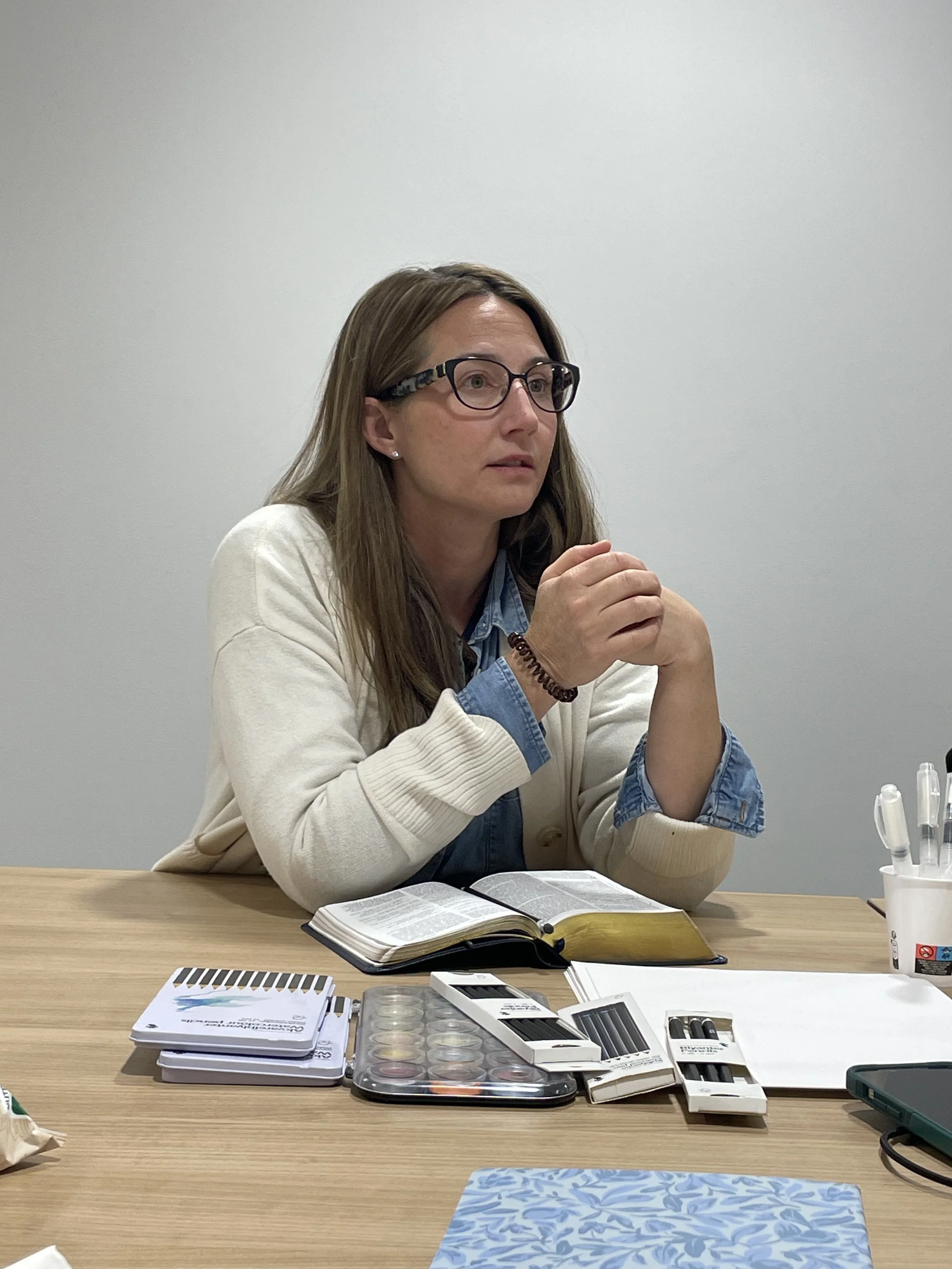 A woman with long brown hair, glasses, wearing a denim shirt under a white cardigan, is sitting at a table with various items, including notebooks, a tray of contact lenses, and eye care products, in a room with a plain white wall.