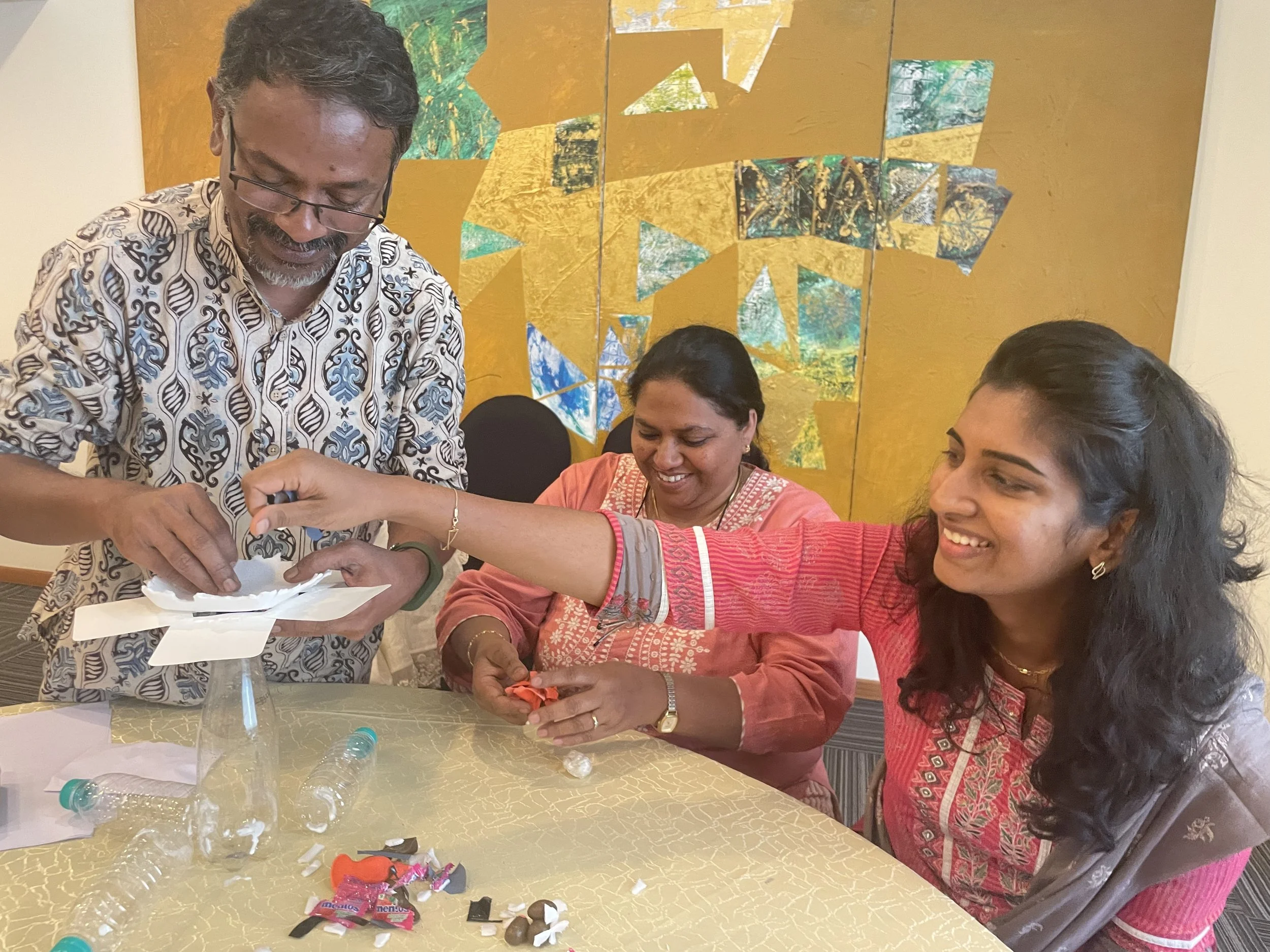 Four people, three women and one man, are seated around a table and engaging in a craft activity. The man, wearing glasses and a patterned shirt, is helping the women. The women are smiling and handling various craft supplies on the table, including shredded paper, plastic bottles, and candy wrappers. The background features a yellow wall with abstract artwork.