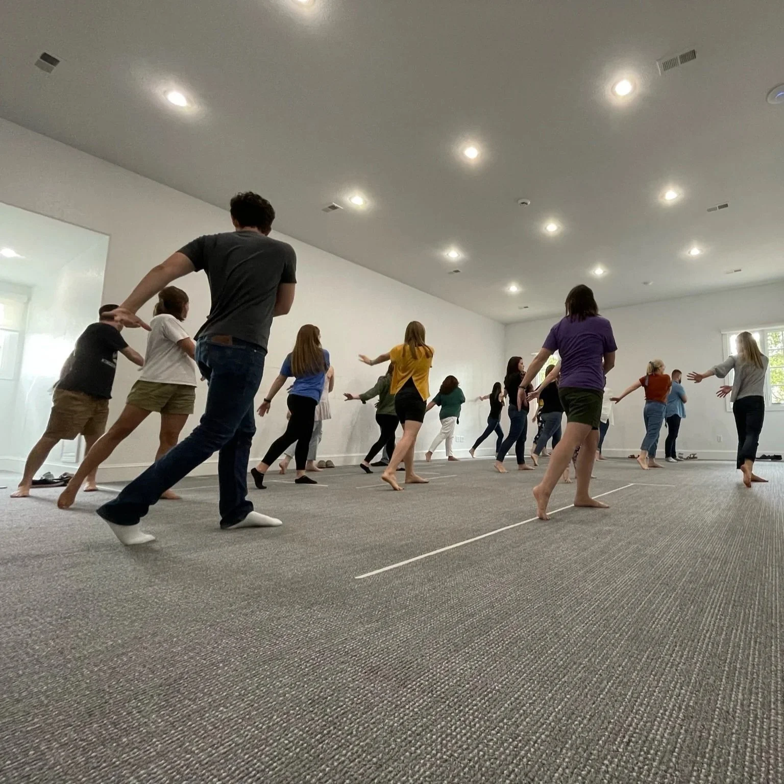 People participating in a dance or exercise class in a large, bright room with white walls and a carpeted floor, some barefoot and some wearing socks.