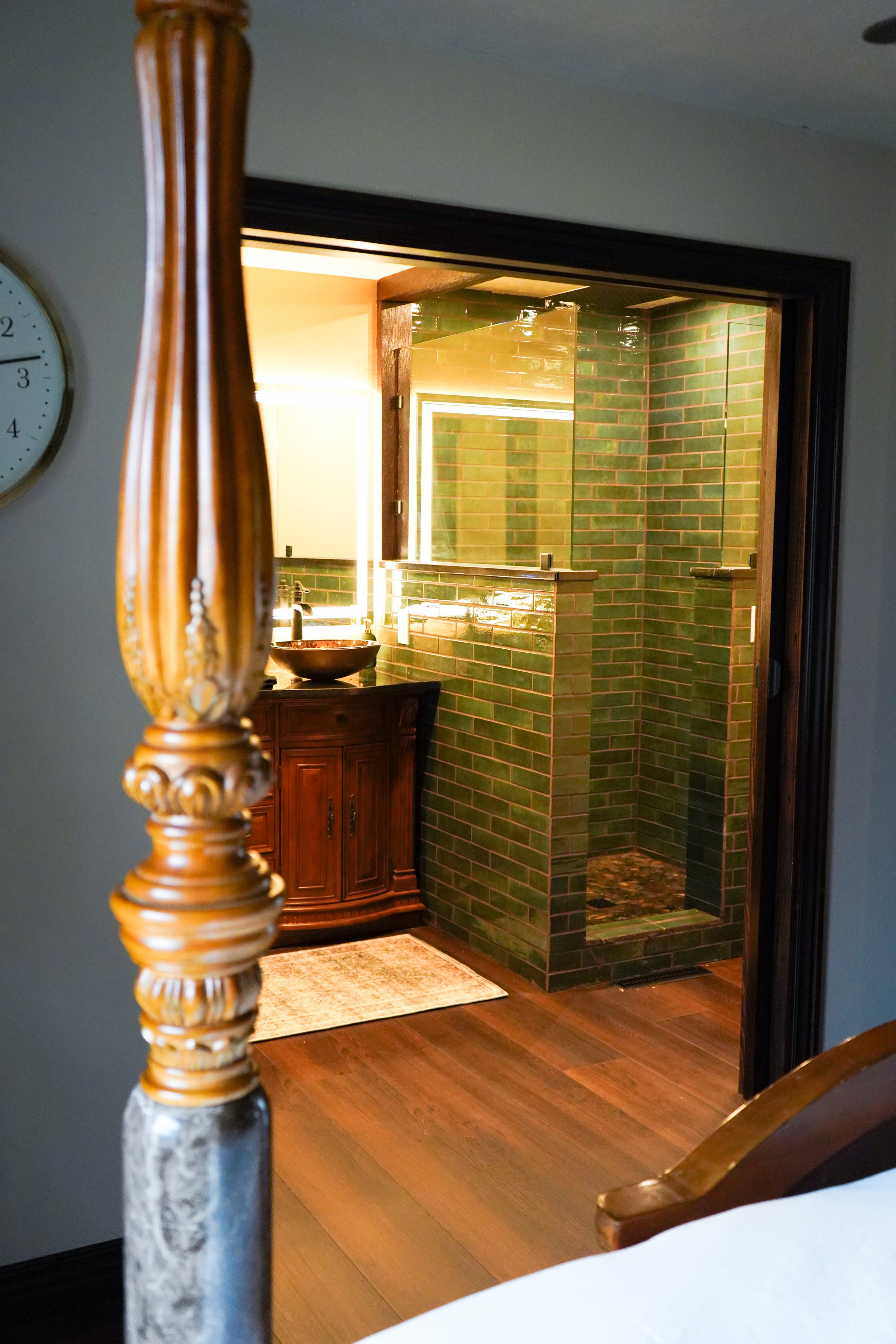 View of a bathroom with green tiled shower area, a wooden vanity with a vessel sink, and a mirror, seen through a bedroom door.