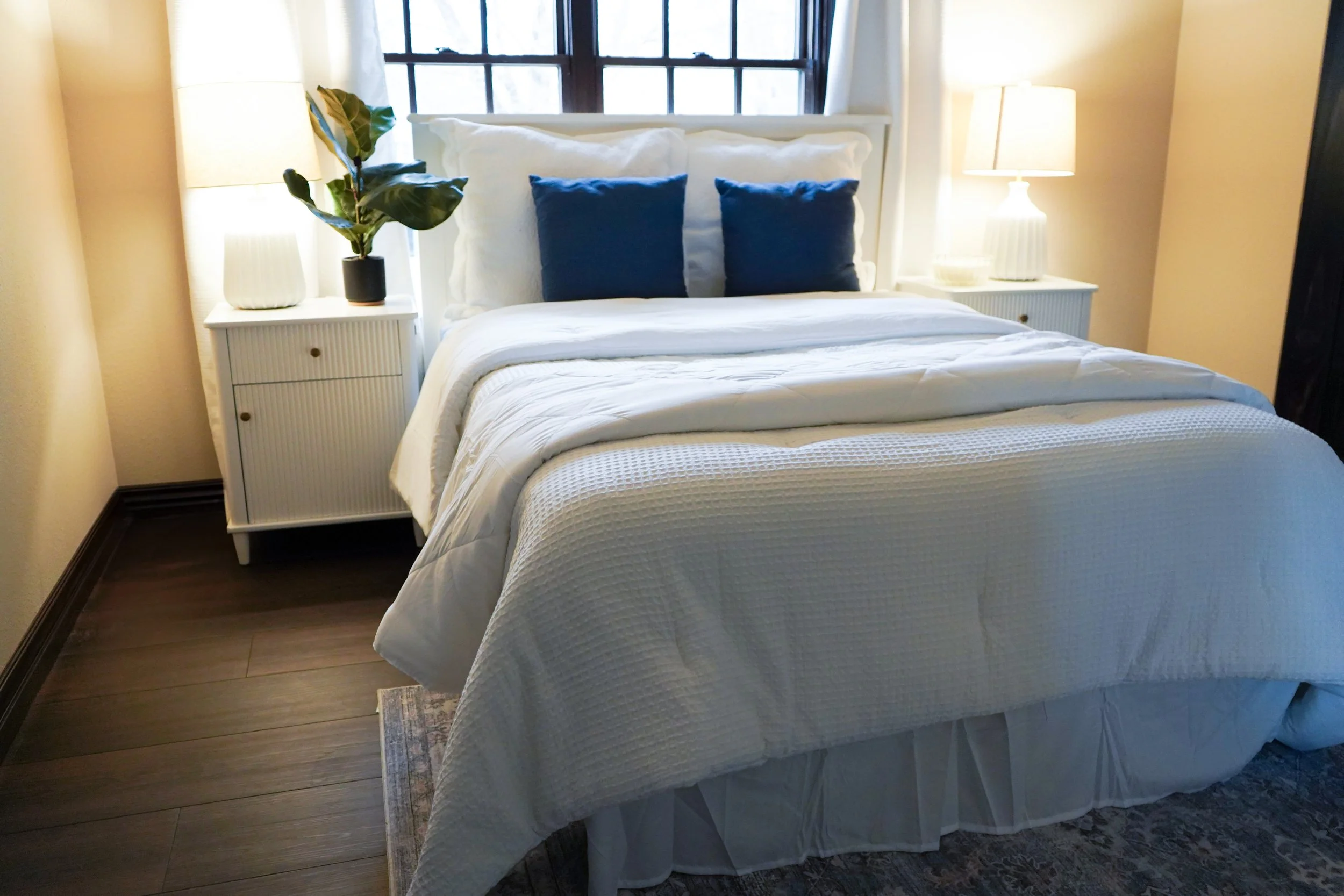 A neatly made bed with white bedding and two dark blue pillows, flanked by white nightstands with lamps and a potted plant on the left nightstand, in a room with dark wooden floors and a window with black framing and white curtains.