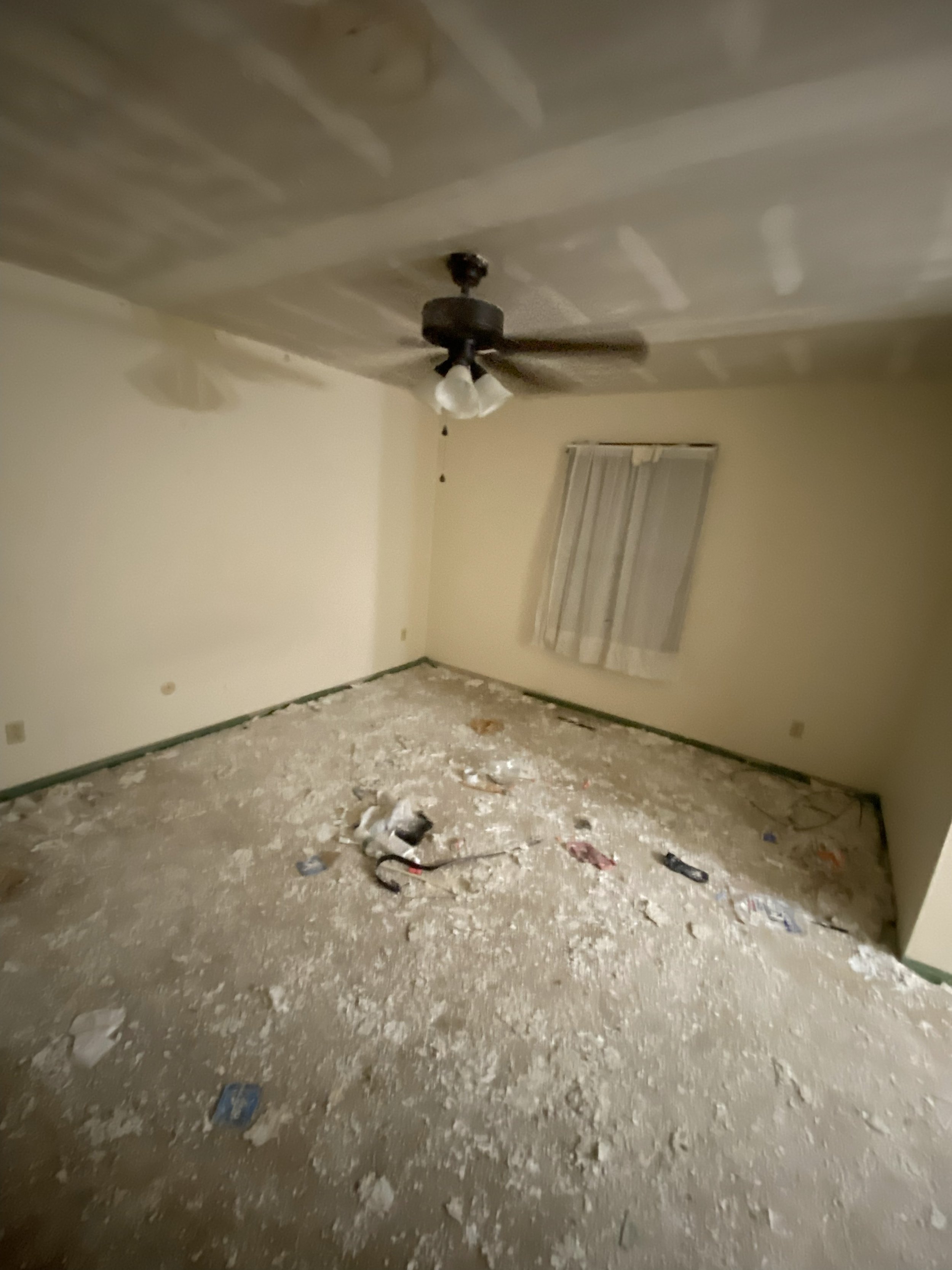 Empty room under renovation with debris on the floor, a ceiling fan, and a window with closed blinds.