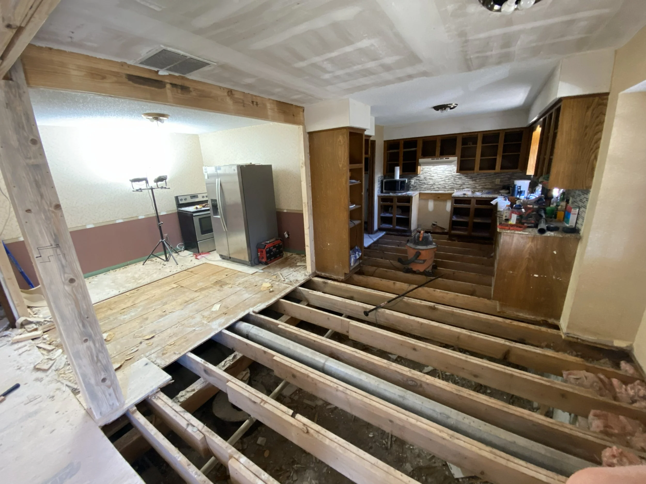 Interior of a home under renovation with exposed floor joists, kitchen cabinets, a microwave, tools, and construction equipment.