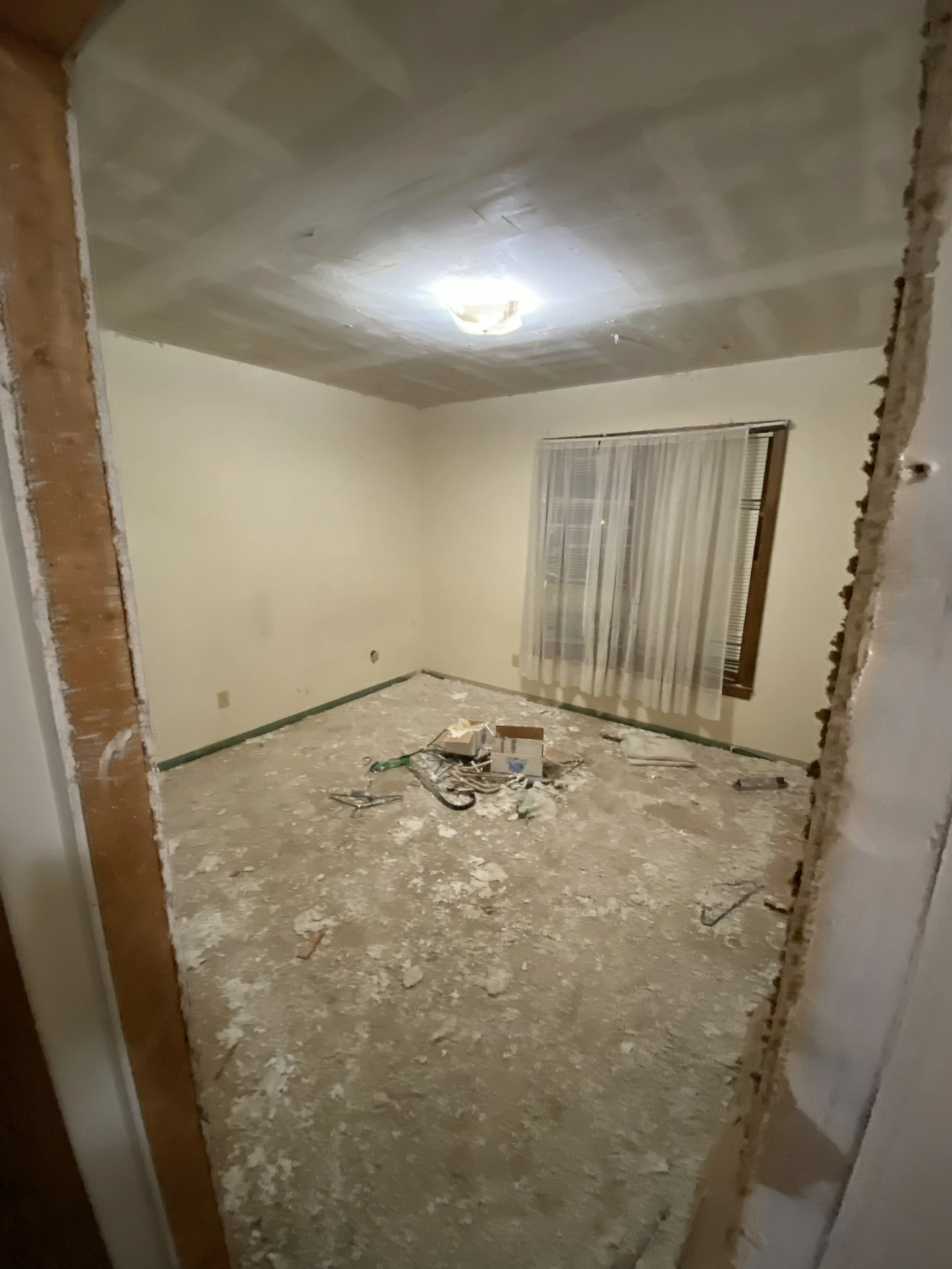 A room under renovation with bare floors, a window with sheer curtains, and construction debris on the floor.
