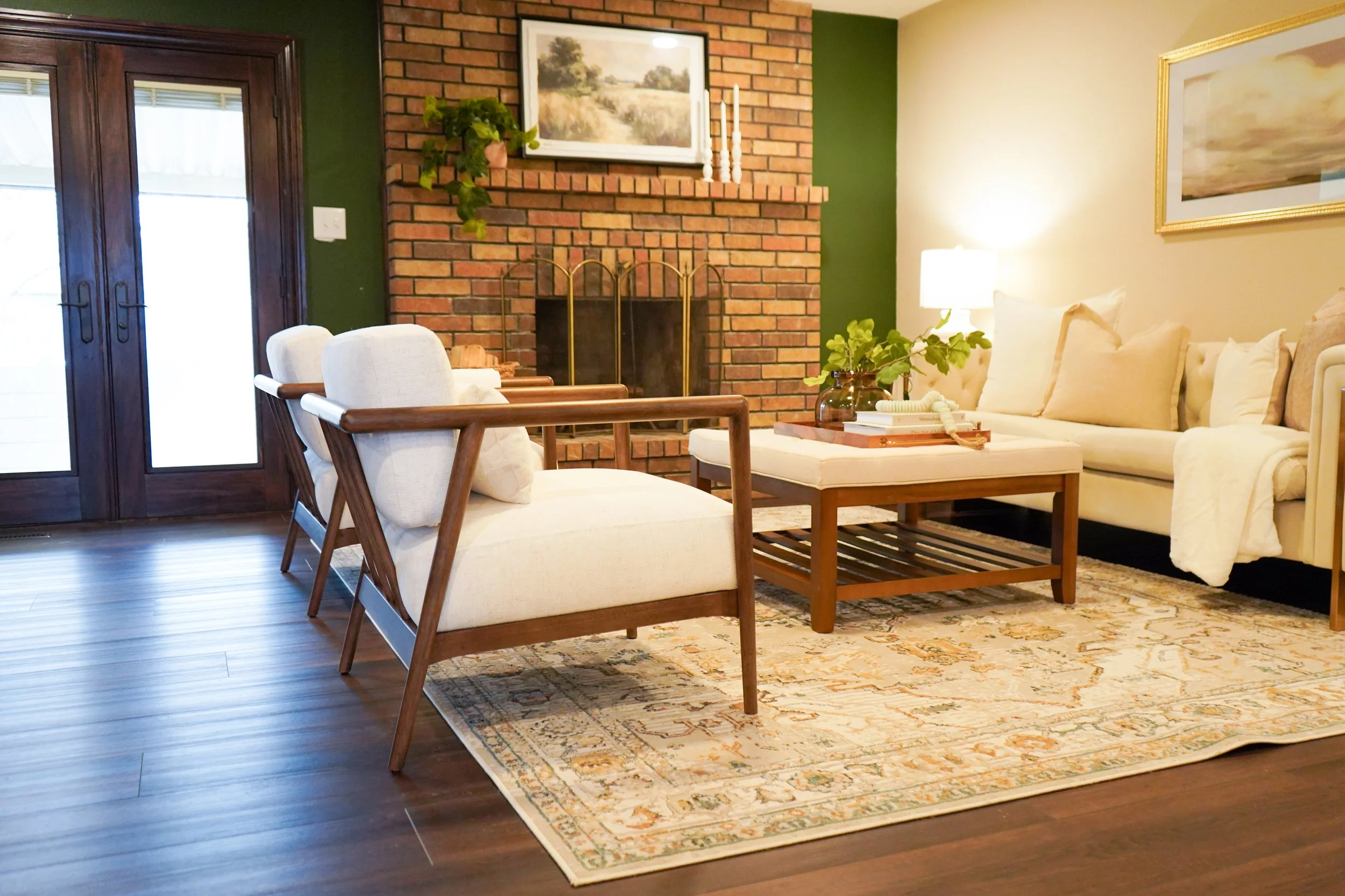 Living room with white sofas, wooden chairs, a brick fireplace, green accent wall, framed artwork, and a patterned area rug.