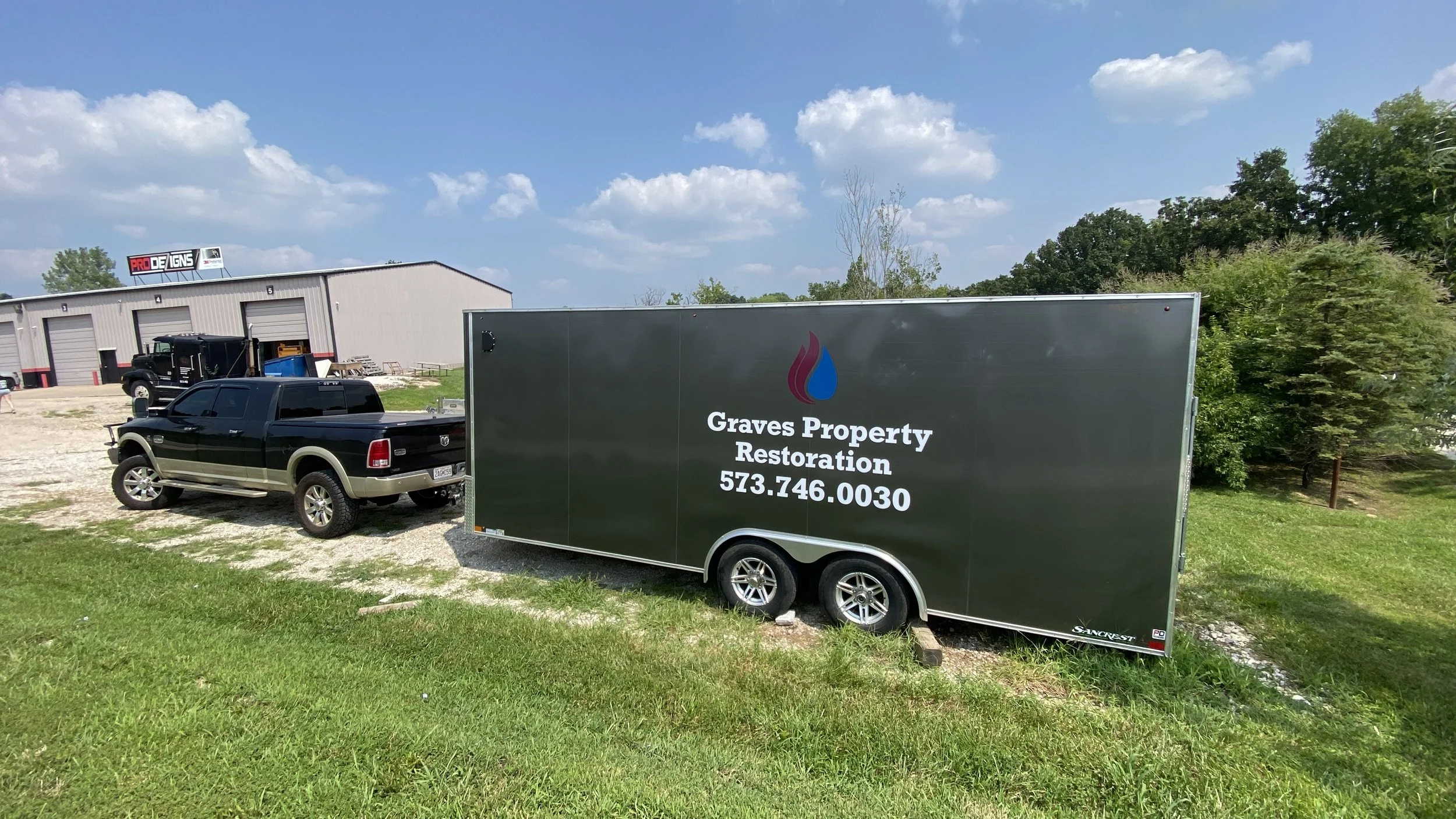 A black trailer with gray text and logo parked on grass next to a black pickup truck, in front of a warehouse with multiple garage doors under a partly cloudy sky.
