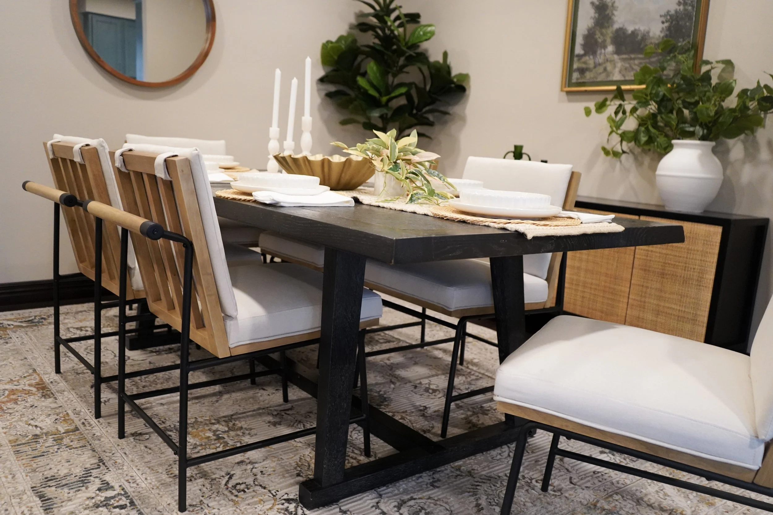Dining room with a large dark wood table set with white plates, napkins, and candles, surrounded by white and wooden chairs, with a patterned rug underneath, and decorated with plants, a mirror, and a framed picture on the wall.