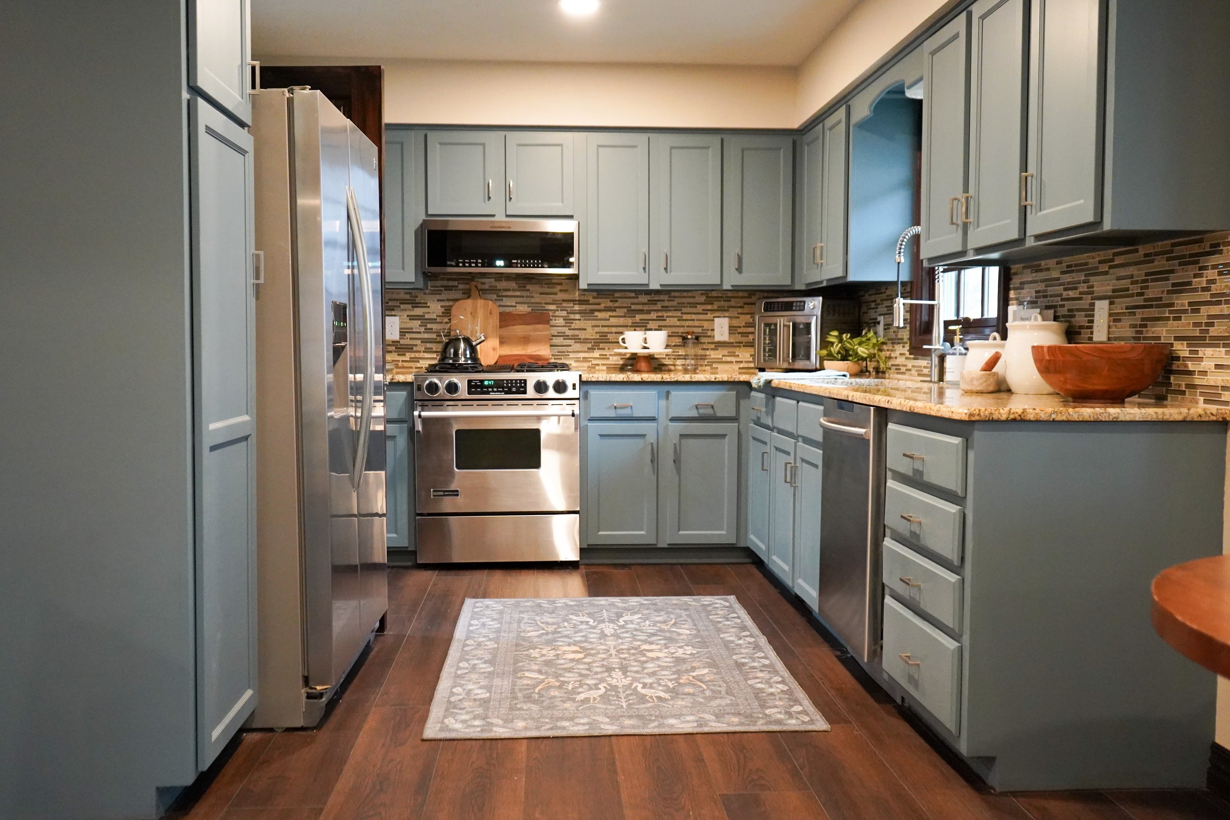 A kitchen with light blue cabinets, stainless steel appliances, a wood countertop, and a decorative tile backsplash. There are various kitchen items such as a wooden bowl, plants, and mugs on the counter. A small rug lies on the dark wood floor.